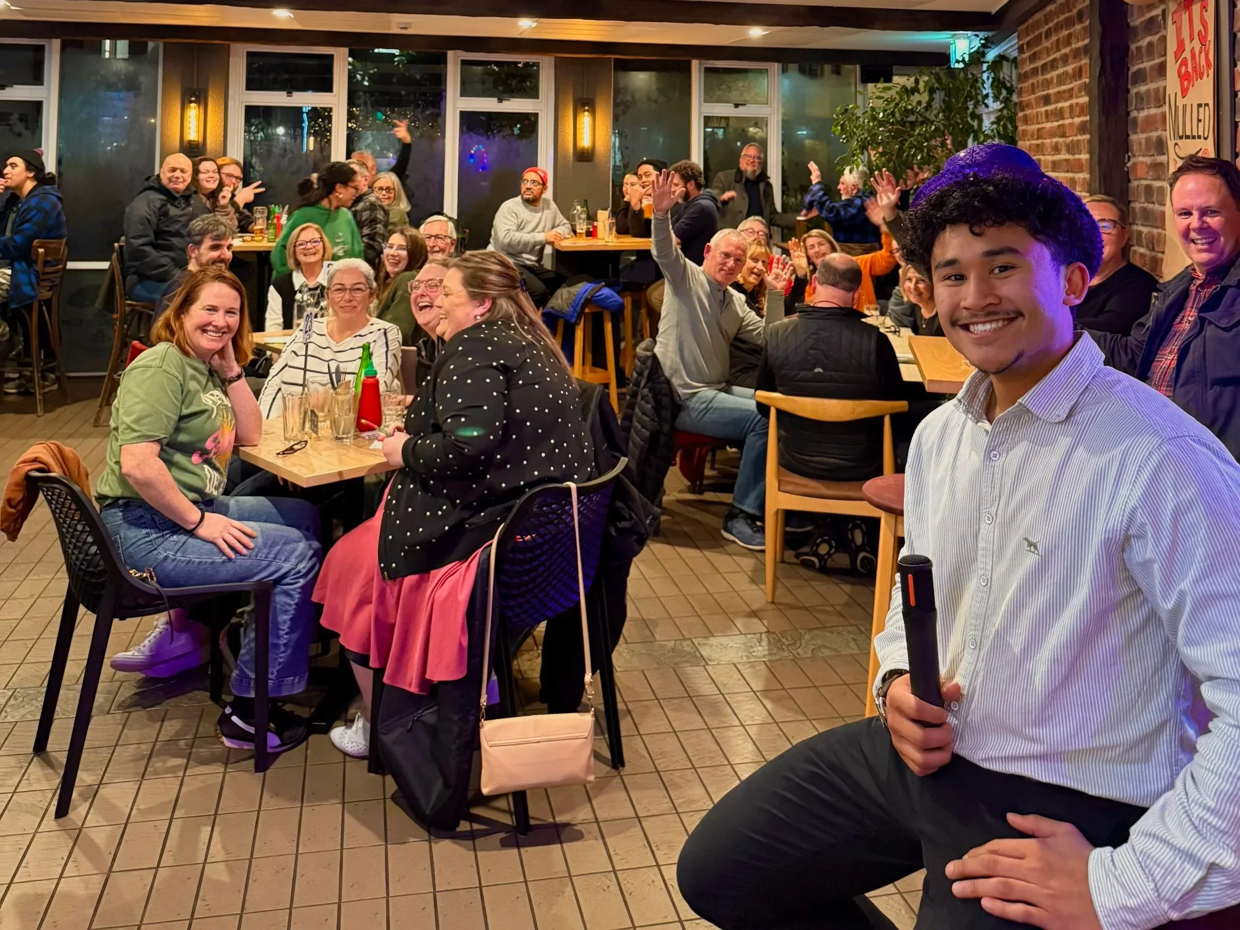 A lively group of people gathered at a restaurant or bar, enjoying drinks and conversation. Many are smiling and some are waving, with a man in the foreground holding a microphone, smiling at the camera.