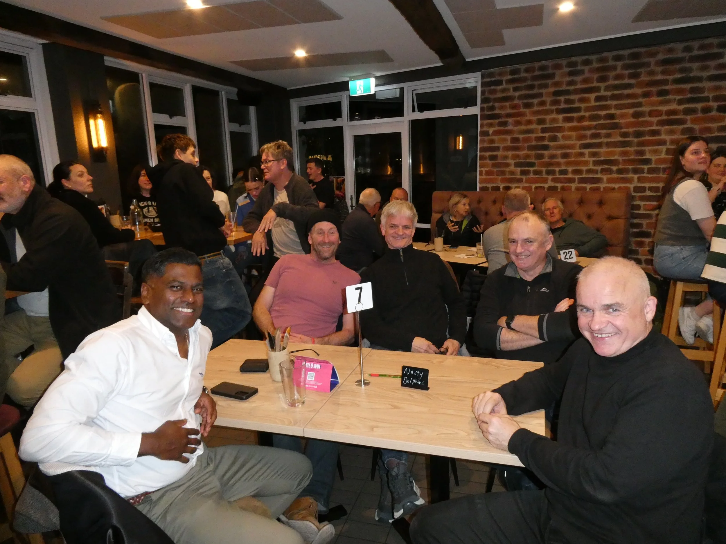 Group of five smiling men sitting at a table in a restaurant or pub, with other people in the background, a brick wall, and large windows behind them.