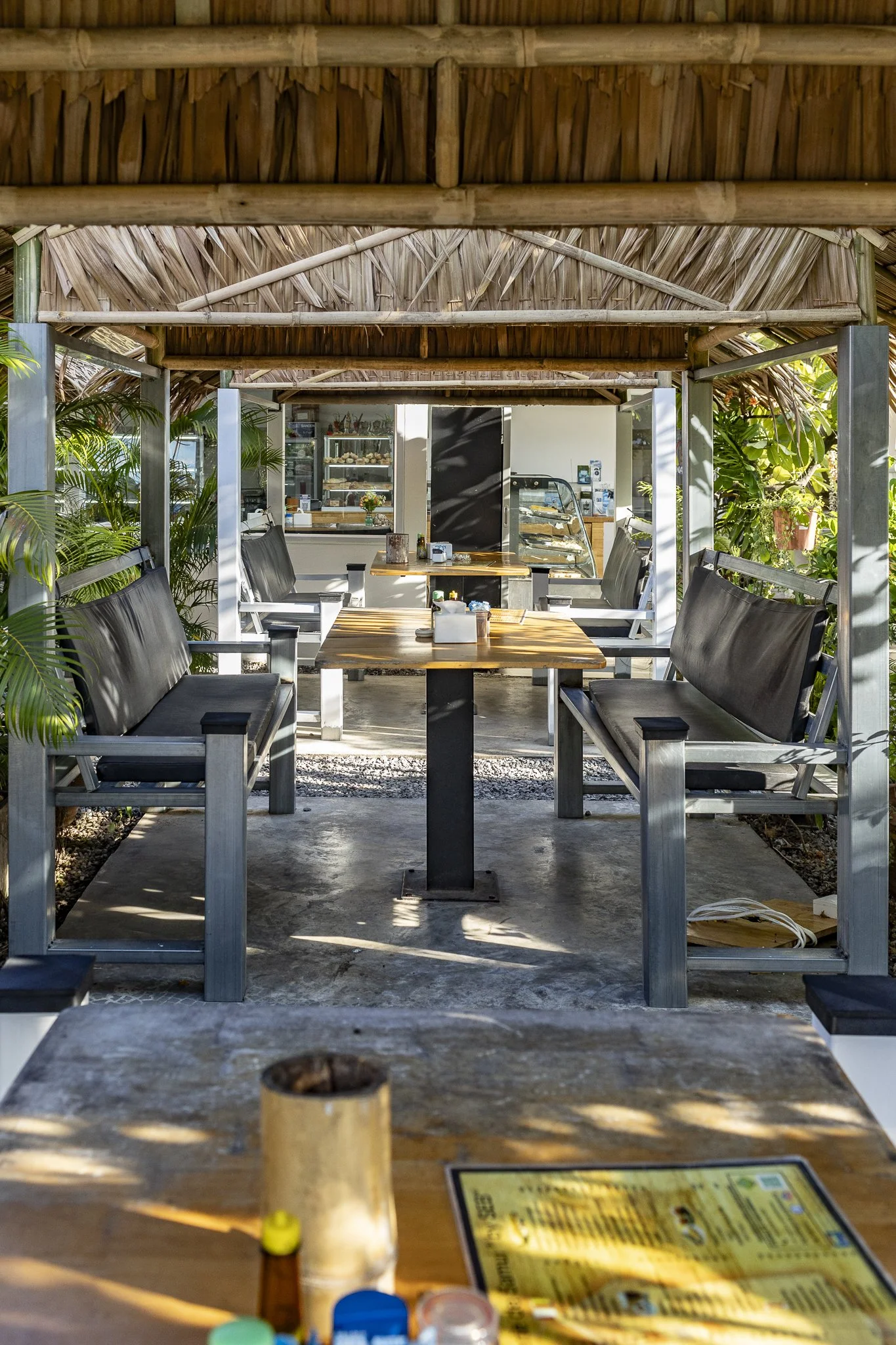 Outdoor seating area with wooden tables and black bench seats under a thatched roof, surrounded by plants, with a view into a cafe in the background.