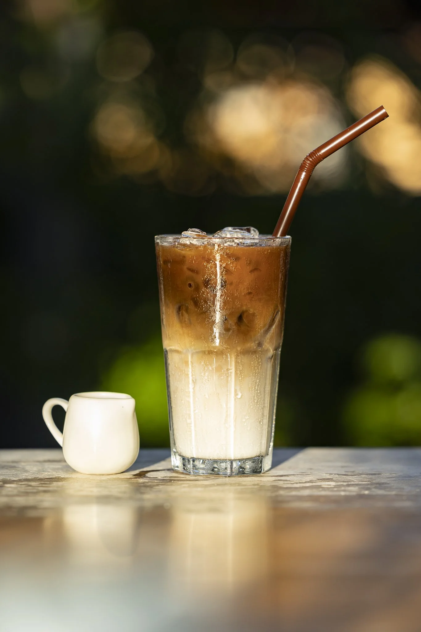 A tall glass of iced coffee with cream at the bottom and coffee on top, with ice cubes and a brown straw, placed on a wooden surface with a small white creamer pitcher beside it, against a blurred outdoor background.