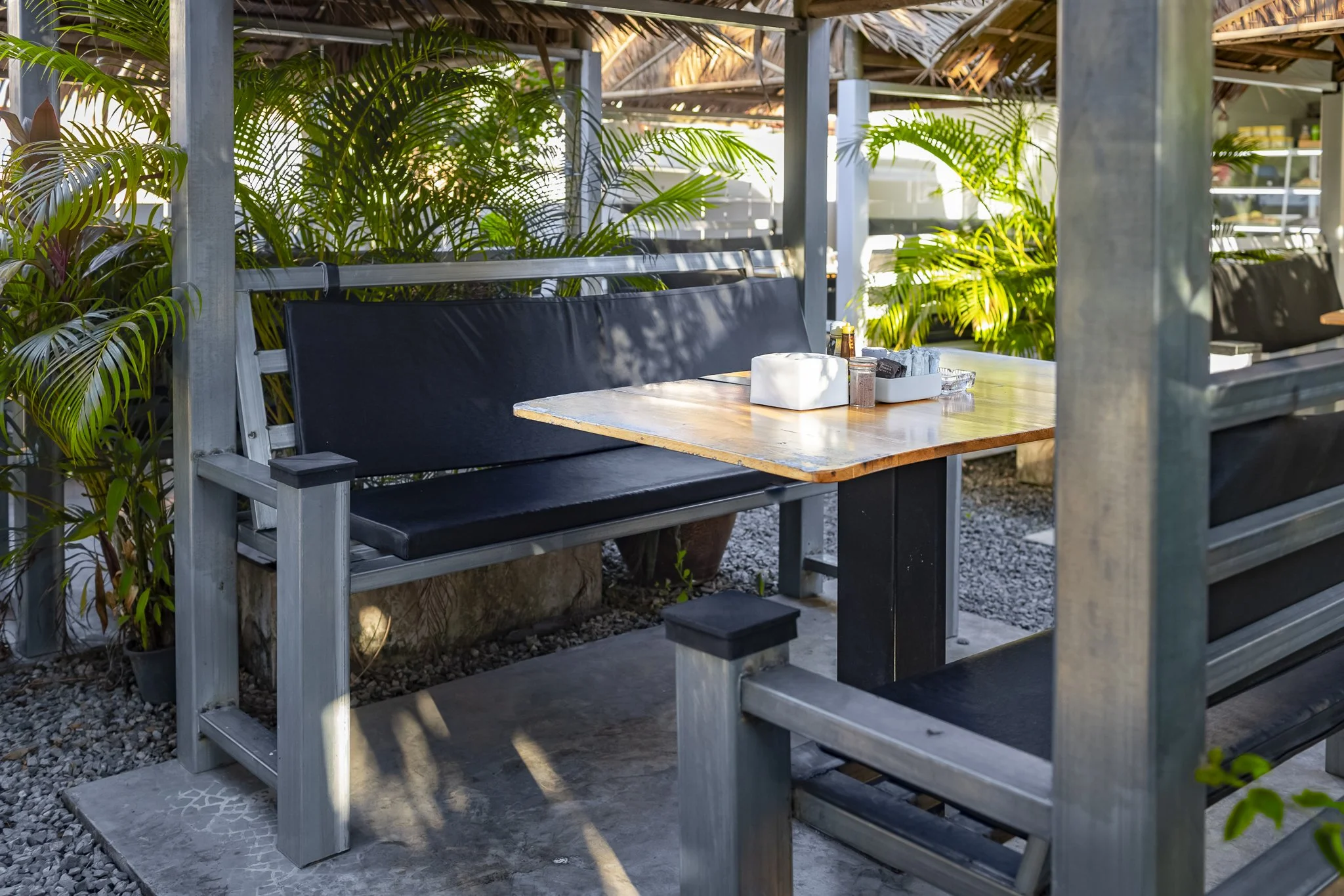 Outdoor dining area with a wooden table, black padded benches, and tropical plants in the background.
