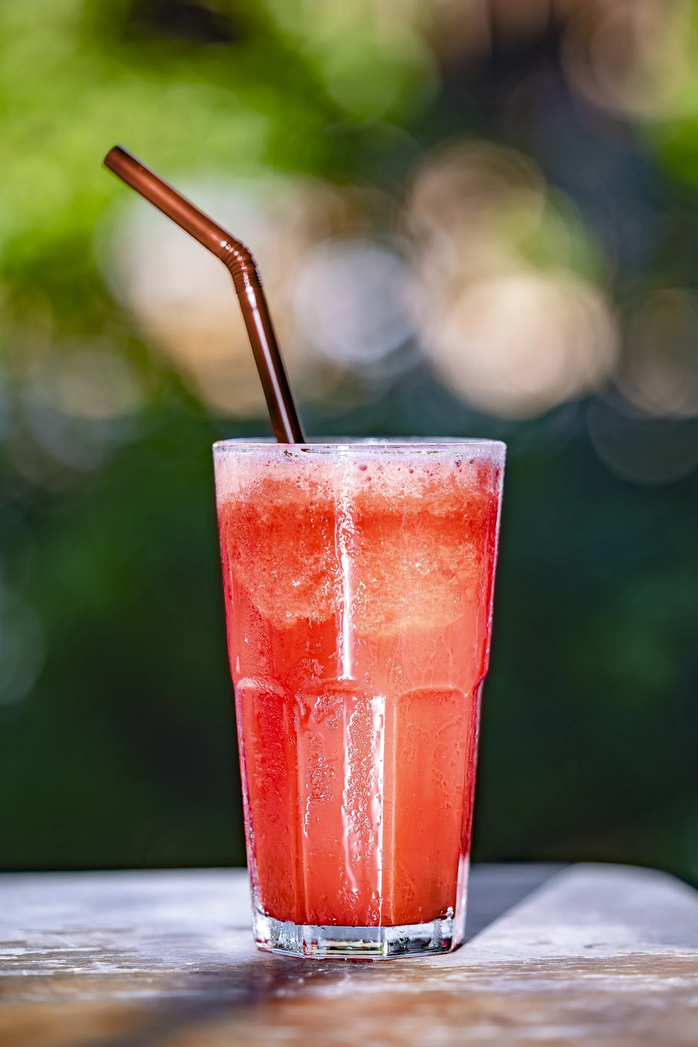A tall glass of red fruit smoothie with a brown straw on a wooden surface, with a blurred green and bokeh background.