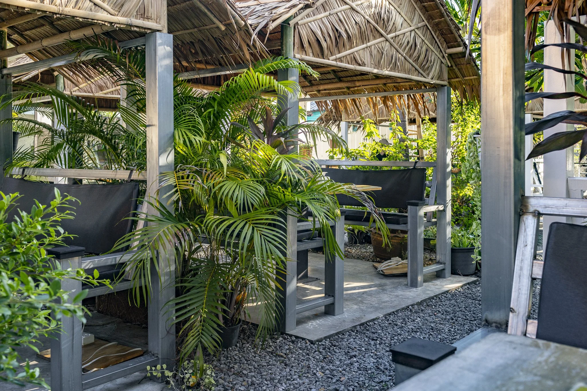 Outdoor seating area with a thatched roof, surrounded by lush green plants and trees, featuring black cushioned benches and a small table on a concrete and gravel ground.