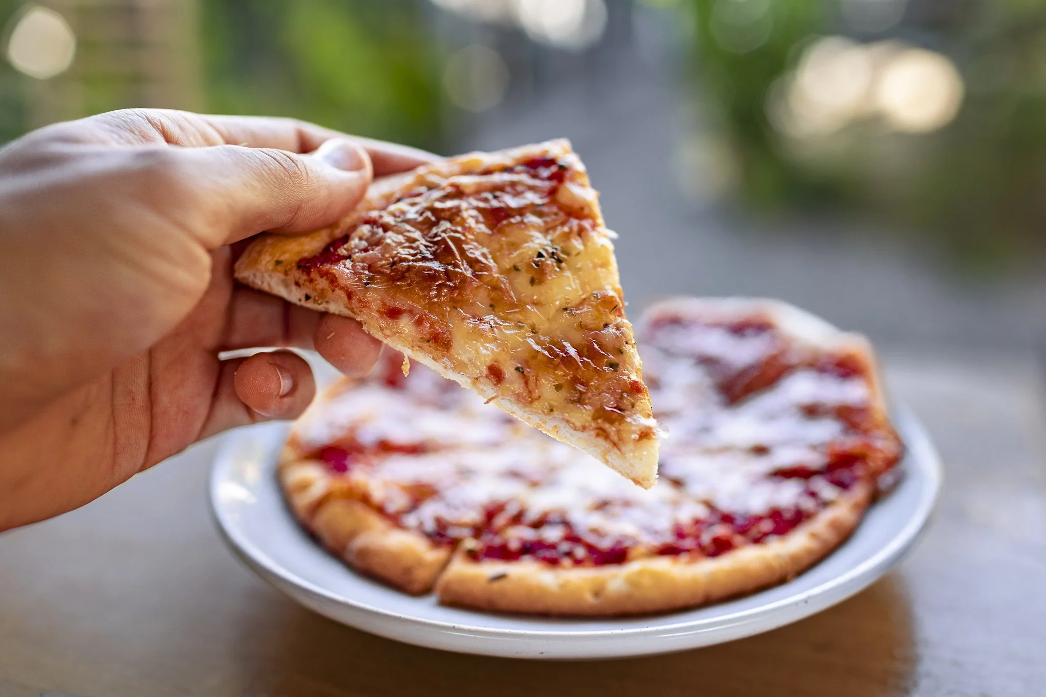 A person holding a slice of cheese pizza above a plate with the remaining pizza, set outdoors on a table.