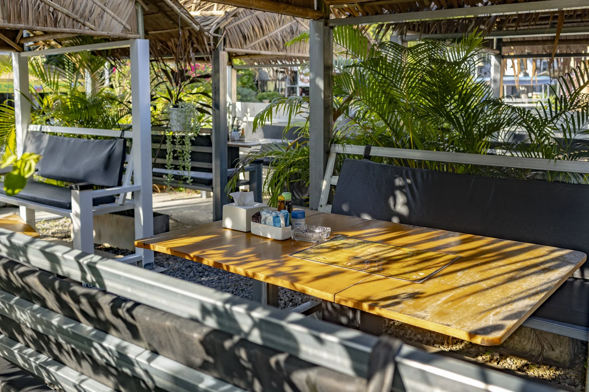 Outdoor restaurant table with condiments, tissues, and sunglasses, shaded by palm fronds, with lush green plants and seating area in the background.