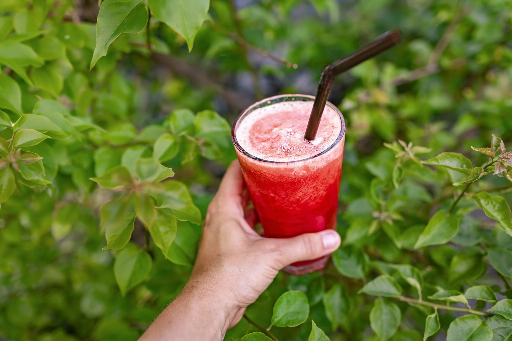 A hand holding a tall glass of pink smoothie with a brown straw, surrounded by green leaves.