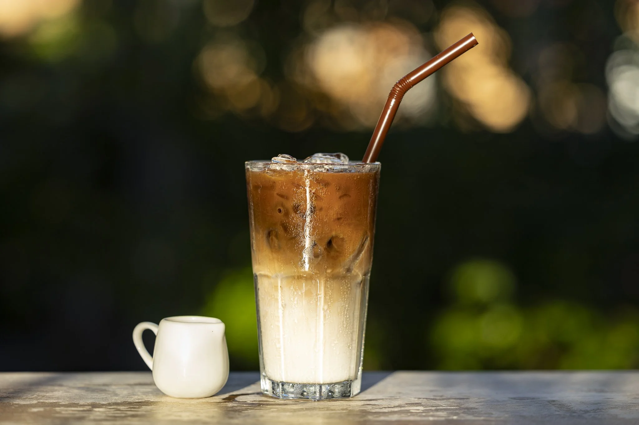 A tall glass of iced coffee with milk, ice cubes, and a brown straw on a wooden surface, with a small white creamer pitcher beside it, outdoors with a blurred natural background.