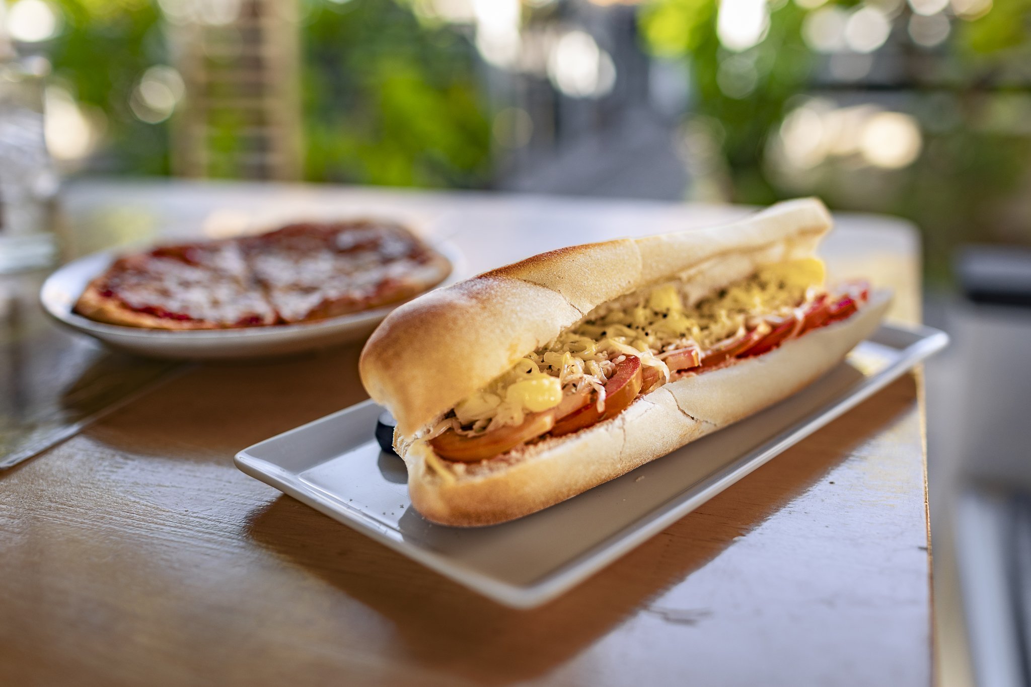 Close-up of a toasted hoagie sandwich with sliced tomatoes, shredded cheese, and possibly chicken, on a white rectangular plate. In the background, there's a plate of pizza on a wooden table, outdoors with blurred greenery.