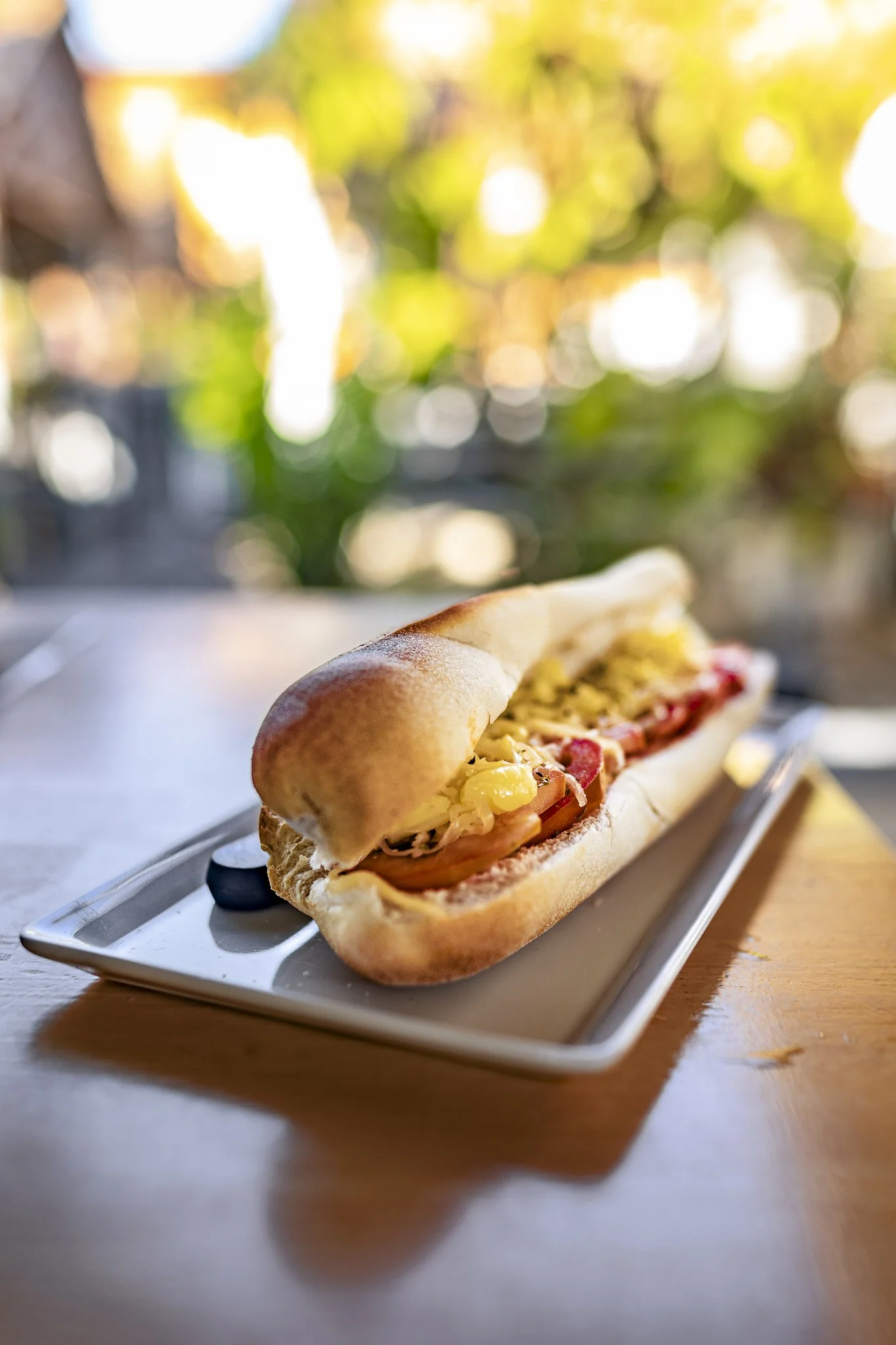 Lemon pepper hot dog in a bun with fried onion rings, on a white rectangular plate on a wooden table, with blurred outdoor background and sunlight.