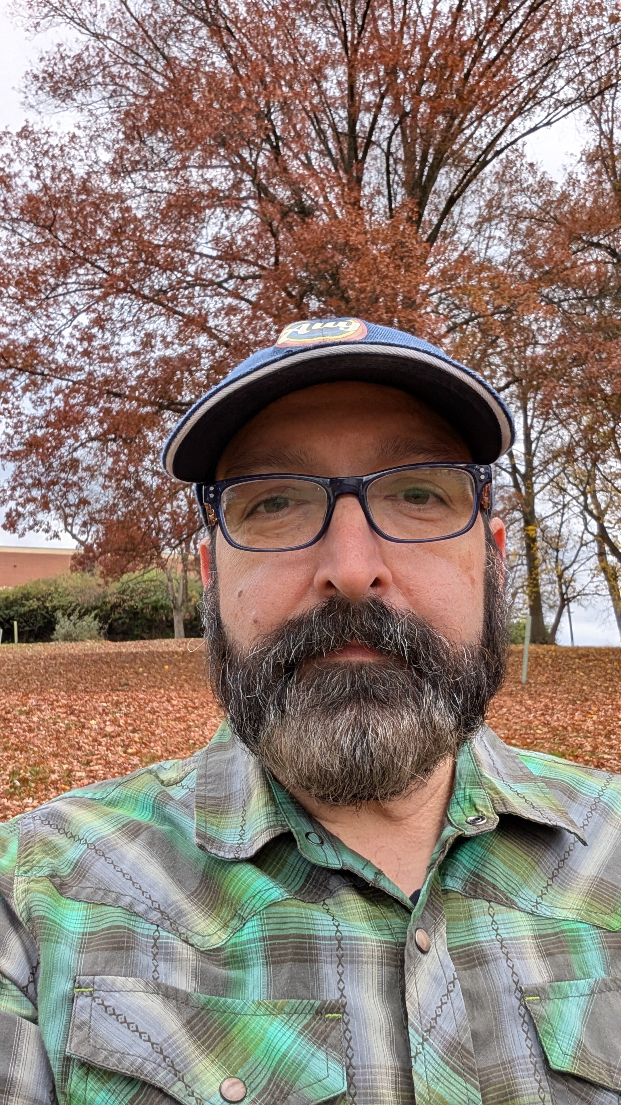 A man with glasses and a beard taking a selfie outdoors with autumn trees in the background.