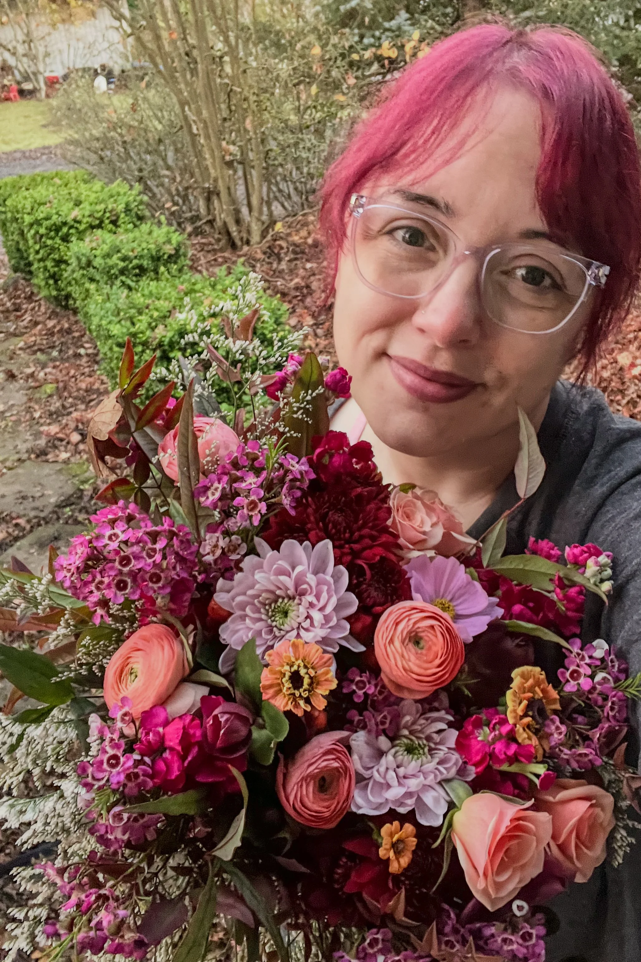 A woman with pink and purple hair and glasses holding a large bouquet of pink, purple, and red flowers outdoors, with bushes and trees in the background.