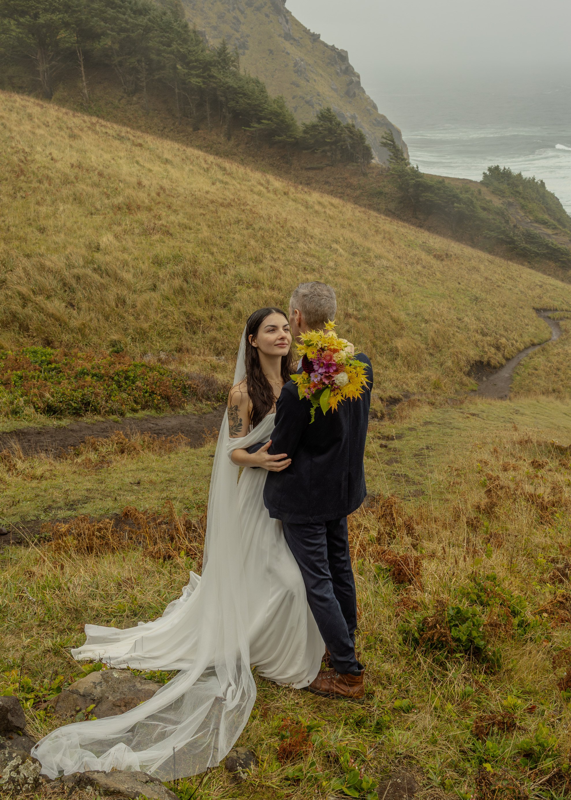 A bride and groom embrace in a scenic outdoor setting with grassy hills, trees, and a distant ocean in the background.