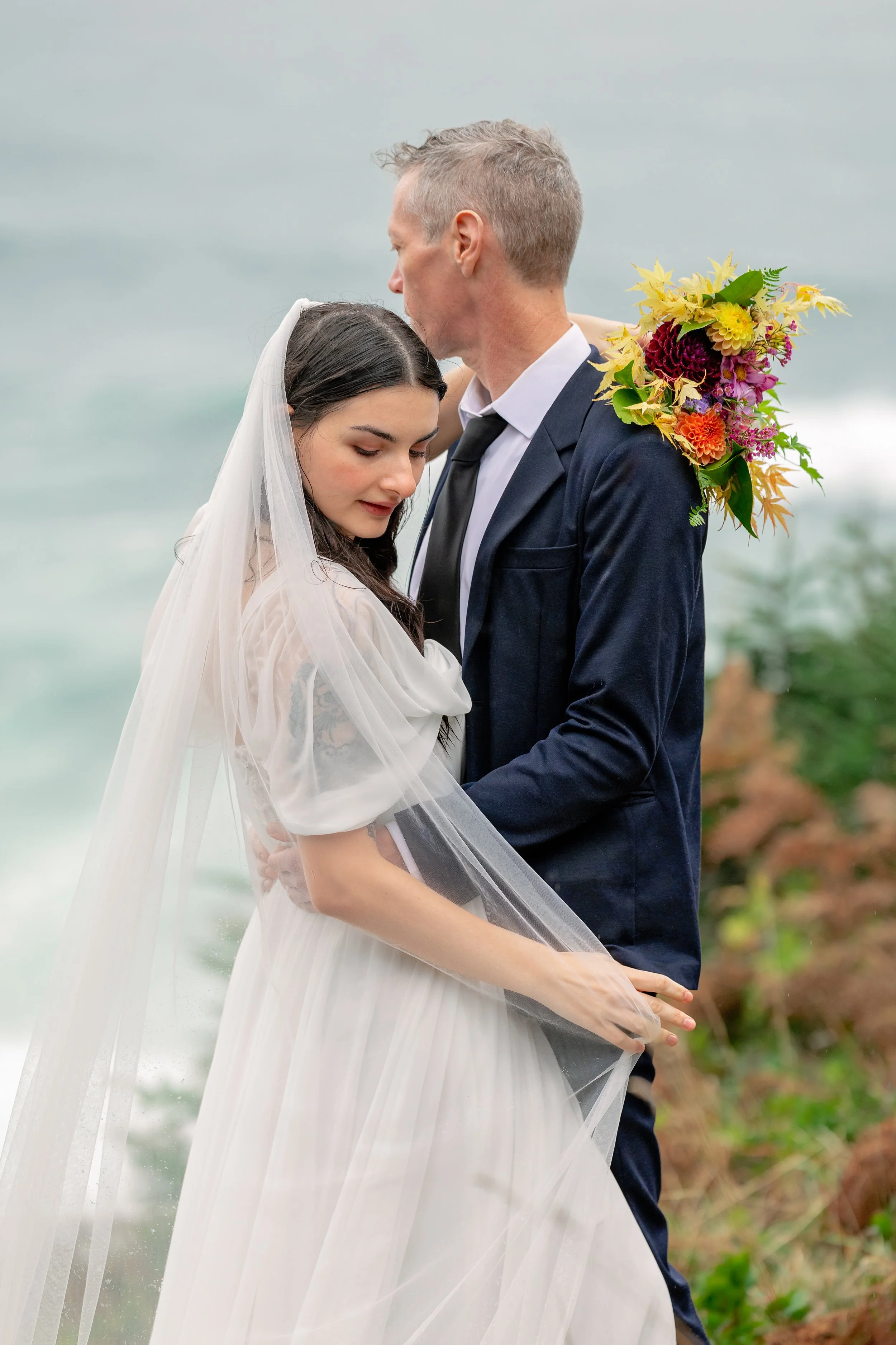 A bride and groom embracing on the beach, with the groom holding a bouquet of colorful flowers over his shoulder. The bride has long dark hair and is wearing a white wedding dress with a veil. The groom has short blond hair and is wearing a dark suit, white shirt, and black tie.