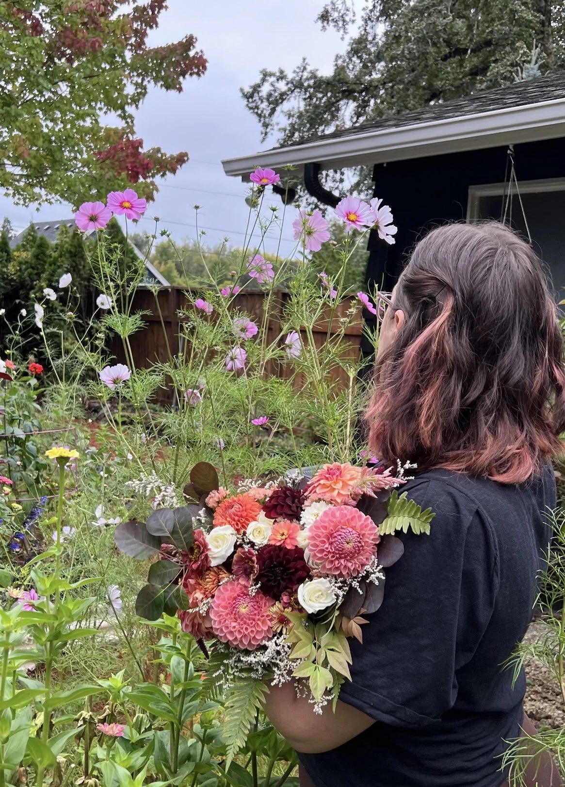 A woman with shoulder-length hair holding a large bouquet of flowers in a garden with various blooming flowers and trees in the background.