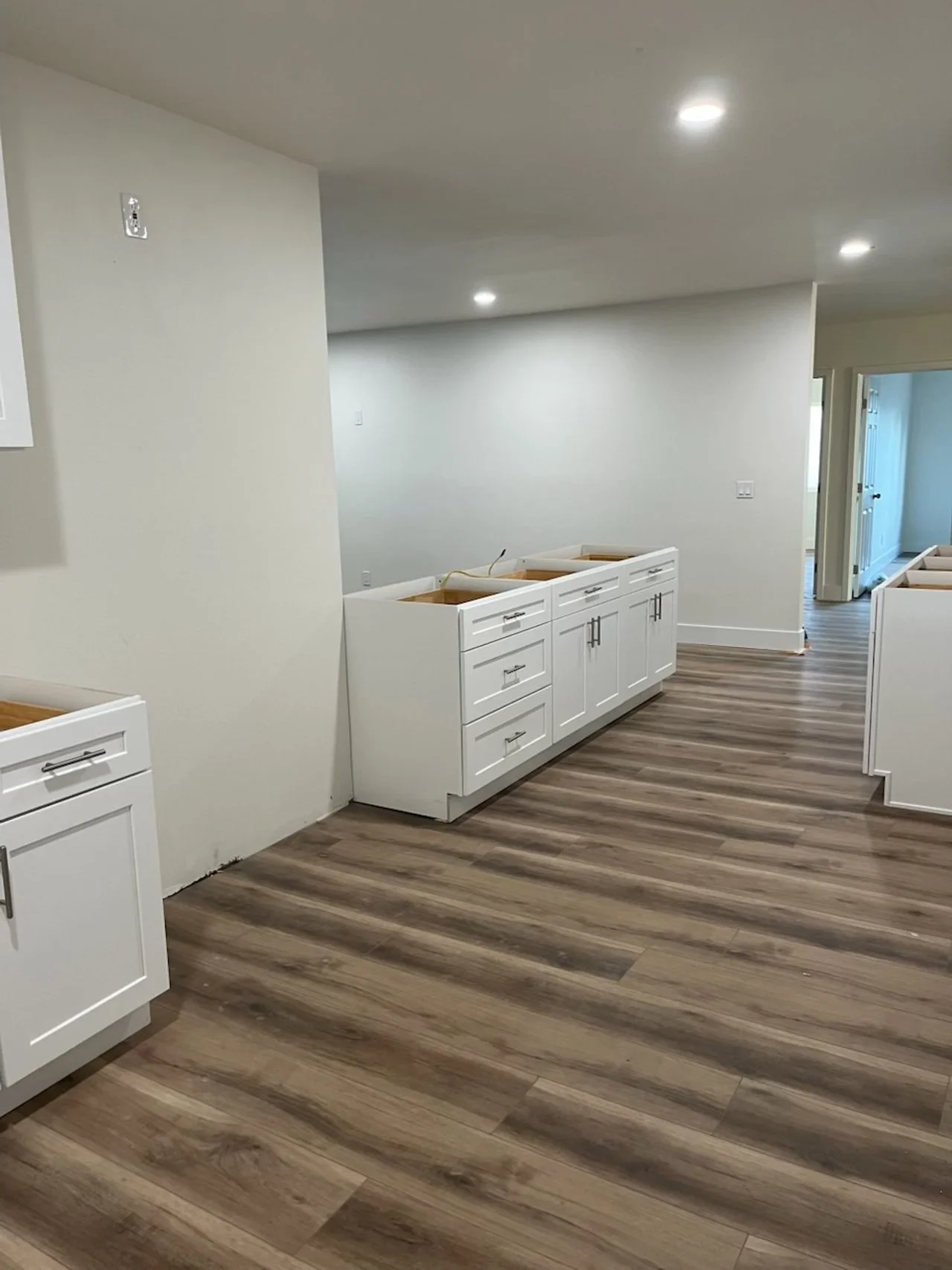Empty kitchen cabinets in a newly constructed room with wood flooring, white walls, and recessed ceiling lights.