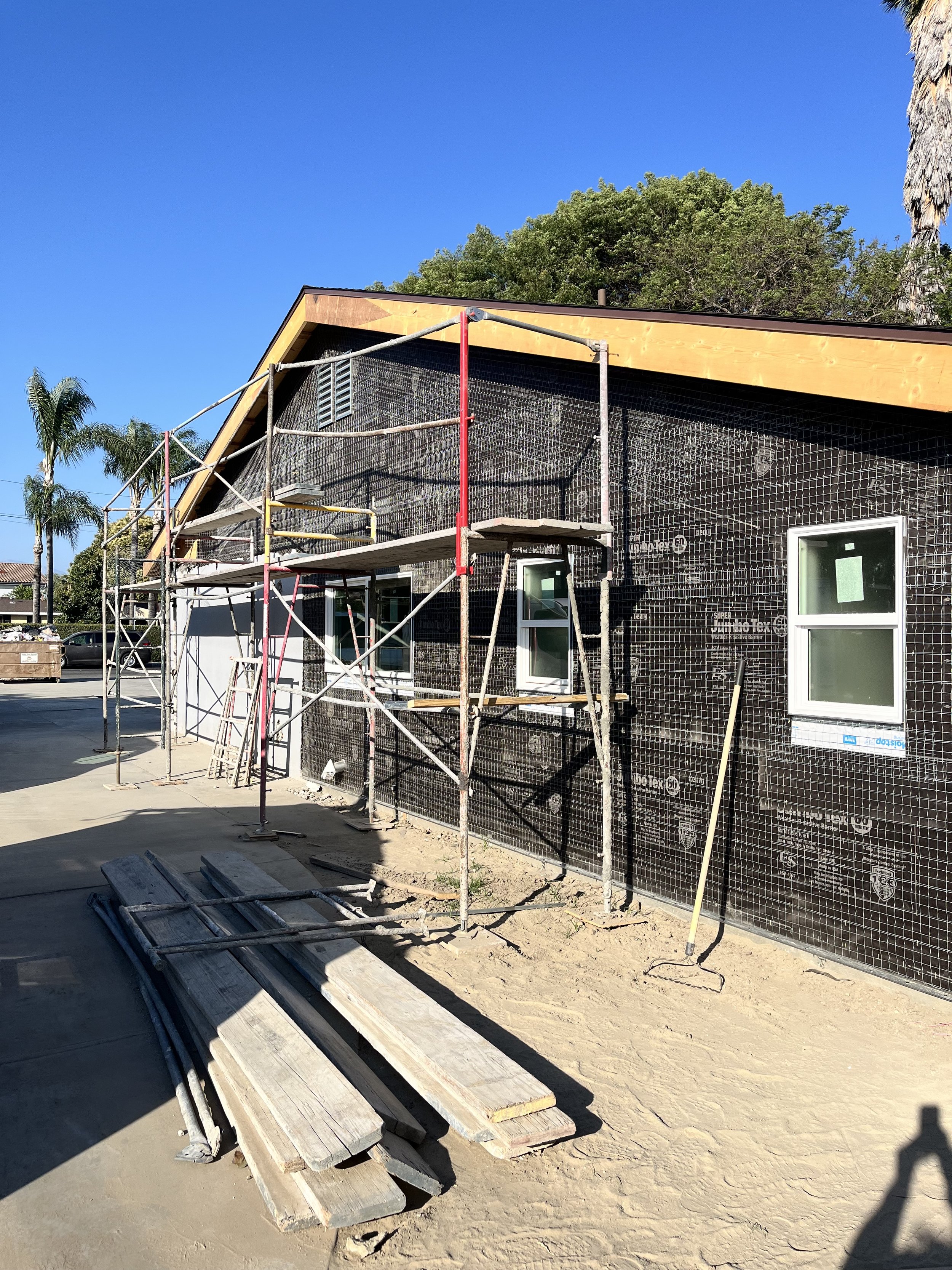 Construction site with scaffolding around a house with black weather-resistant barrier, two white windows, and palm trees in the background under a clear blue sky.