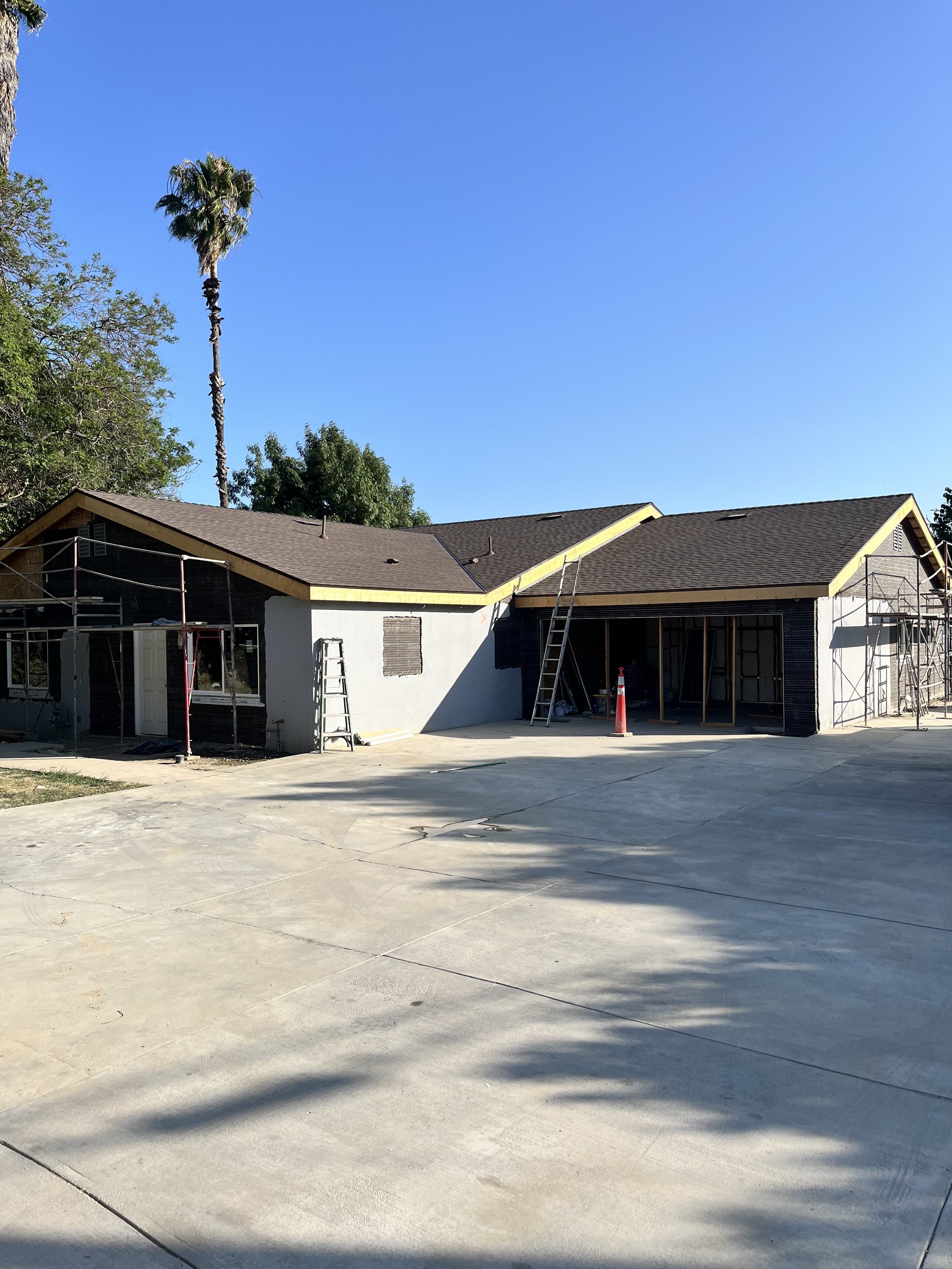 Construction site of a house with scaffolding, ladders, and a cone, with a large driveway and a tall palm tree in the background under a blue sky.