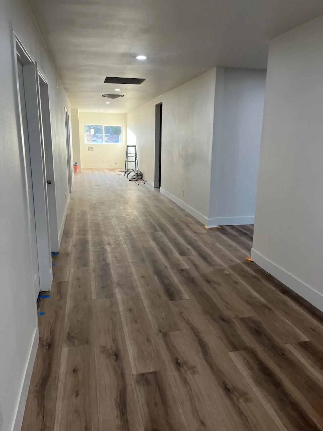 An interior hallway of a house under renovation, with light wood flooring, white walls, and a ladder near the window at the end.