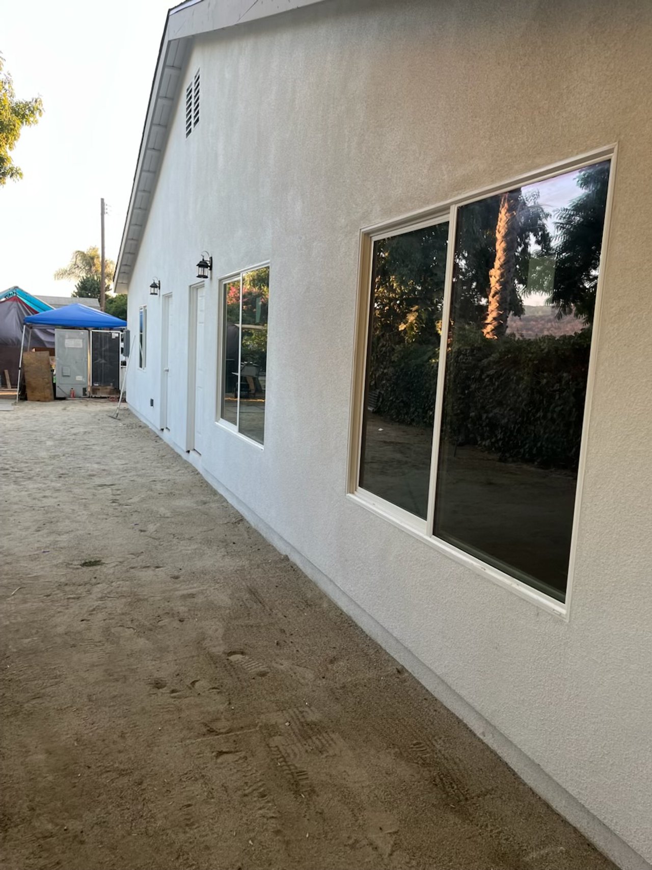 Side view of a house with white stucco exterior and large windows, outdoors with dirt ground and some construction tents and materials in the background.
