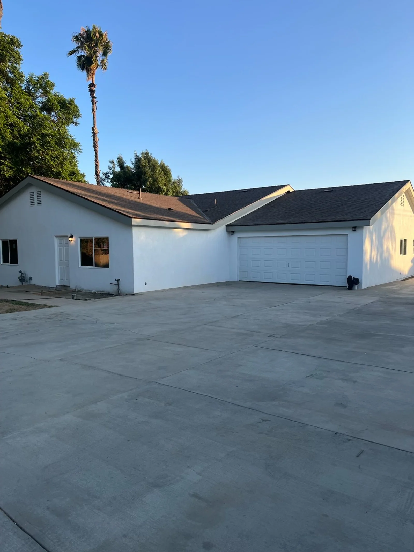 White single-story house with a two-car garage, situated on a concrete driveway, with a tall palm tree and another tree in the background under a clear blue sky.