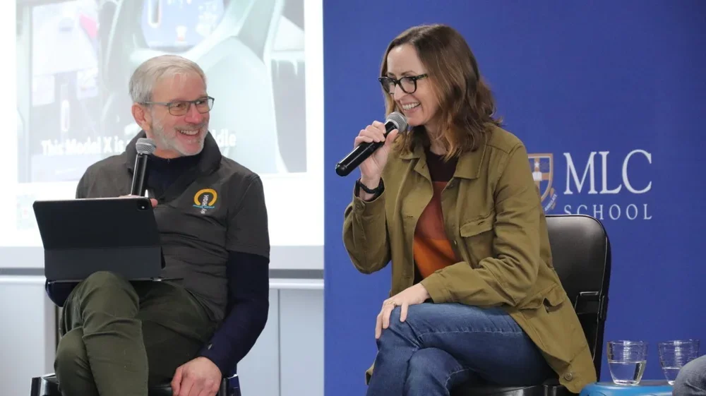 Two people, a man and a woman, are sitting on stage with microphones in their hands, smiling and engaged in conversation. There is a presentation screen behind them and a blue banner with the words 'MLC School' on it.