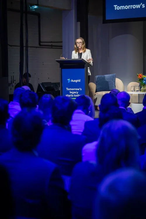 A woman standing at a podium giving a presentation to an audience, with a large screen displaying the word "Tomorrow's" and a sign on the podium with the Ausgrid logo and the phrase "Tomorrow's legacy starts today." The audience is seated and facing the speaker, and there are chairs and a table with flowers on stage.