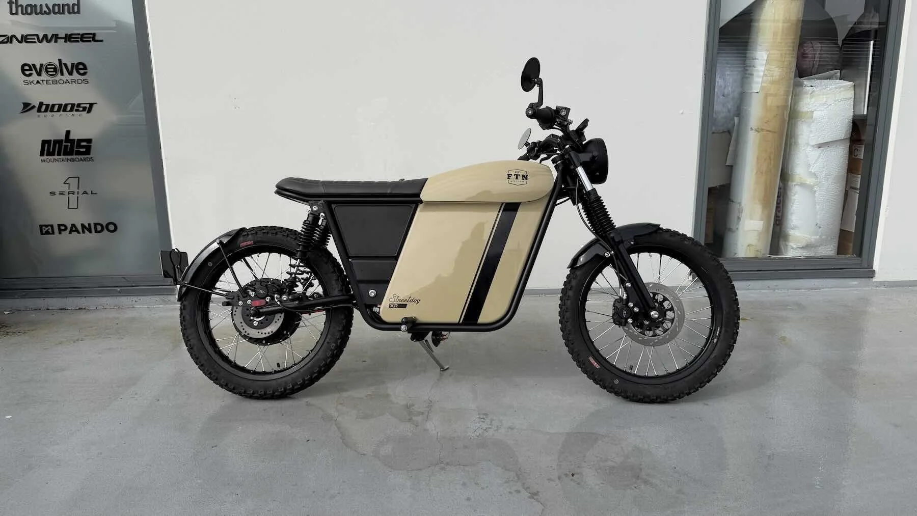 A beige electric bicycle with black accents and off-road tires parked indoors on a concrete floor, against a white wall, with a storage cabinet filled with rolls of material in the background.