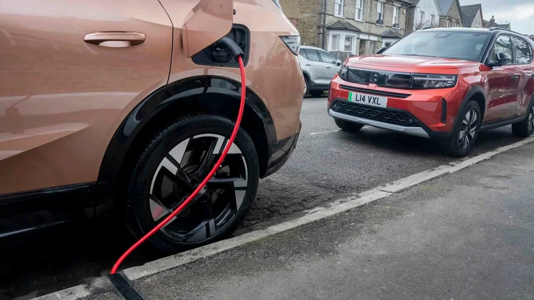 An electric vehicle charging station where a brown car is plugged in, with a red charging cable connected to it, parked next to a red car on a city street.