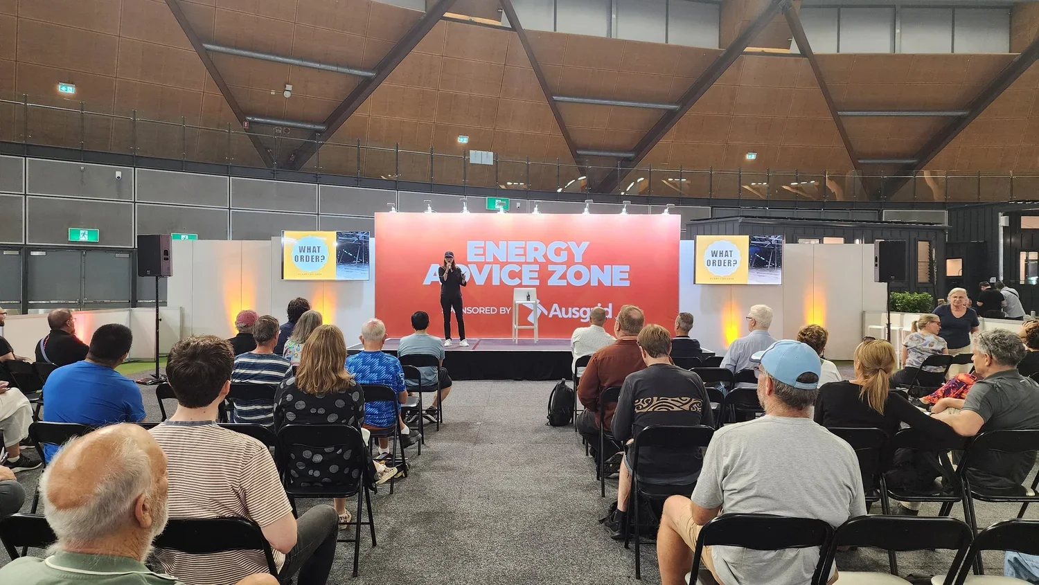 A speaker presenting at the Energy Advice Zone event, with a large red backdrop and audience seated facing the stage in an auditorium.