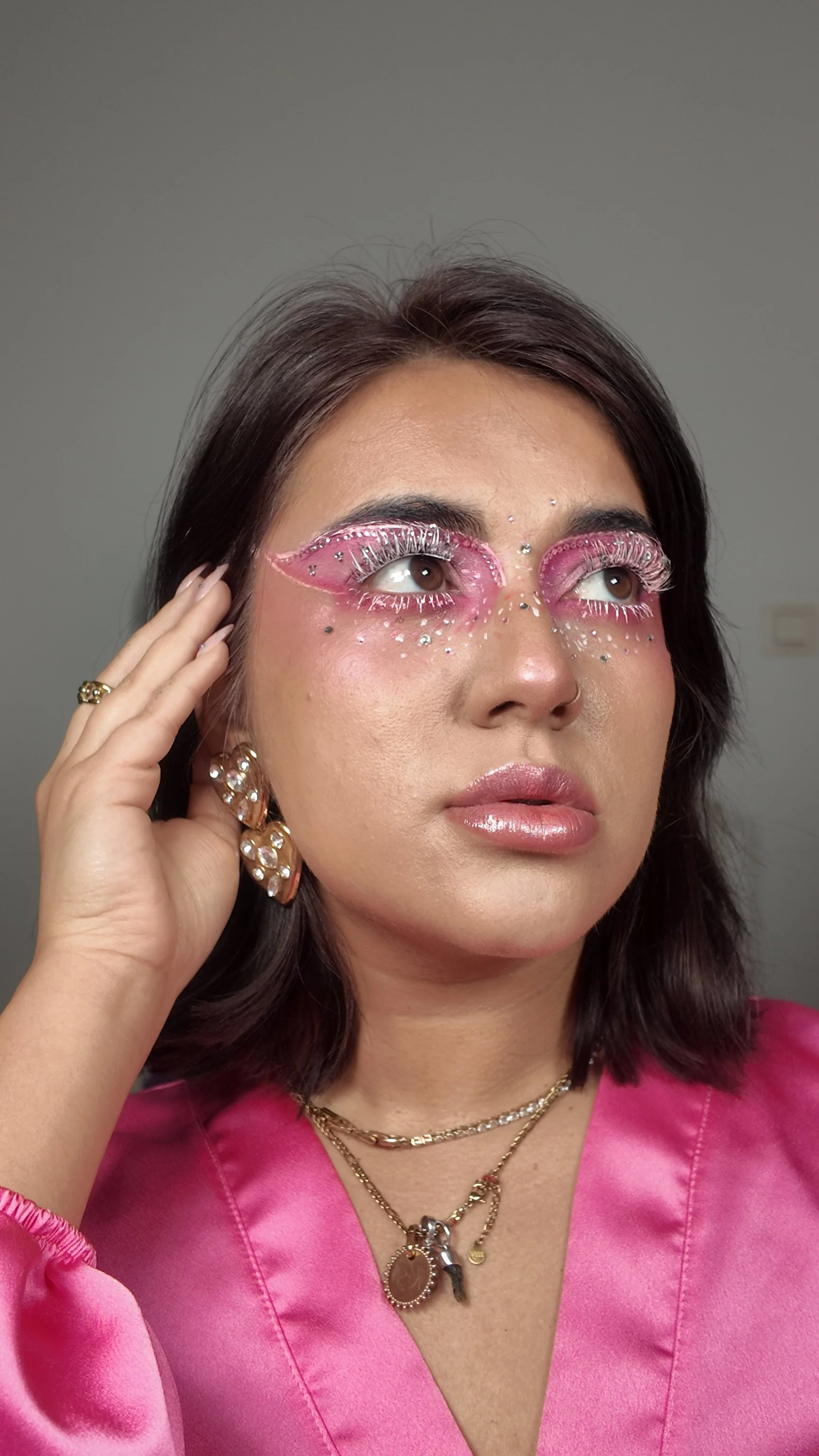 A woman with dark hair wearing bold pink and white eye makeup with rhinestones and feathered eyelashes, dressed in a pink satin top, accessorized with layered necklaces and pearl earrings.