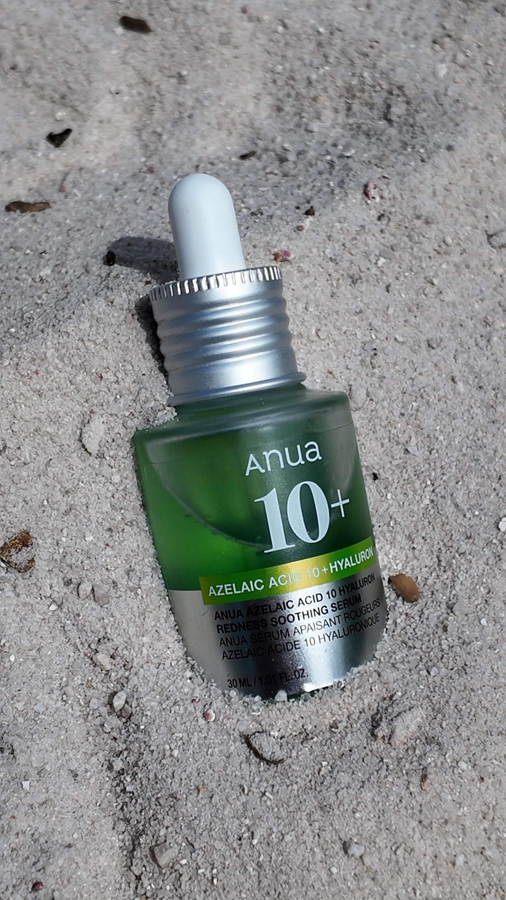 A small green bottle of Anua 10+ Azelaic Acid and Hyaluronic Serum lying on sandy ground.