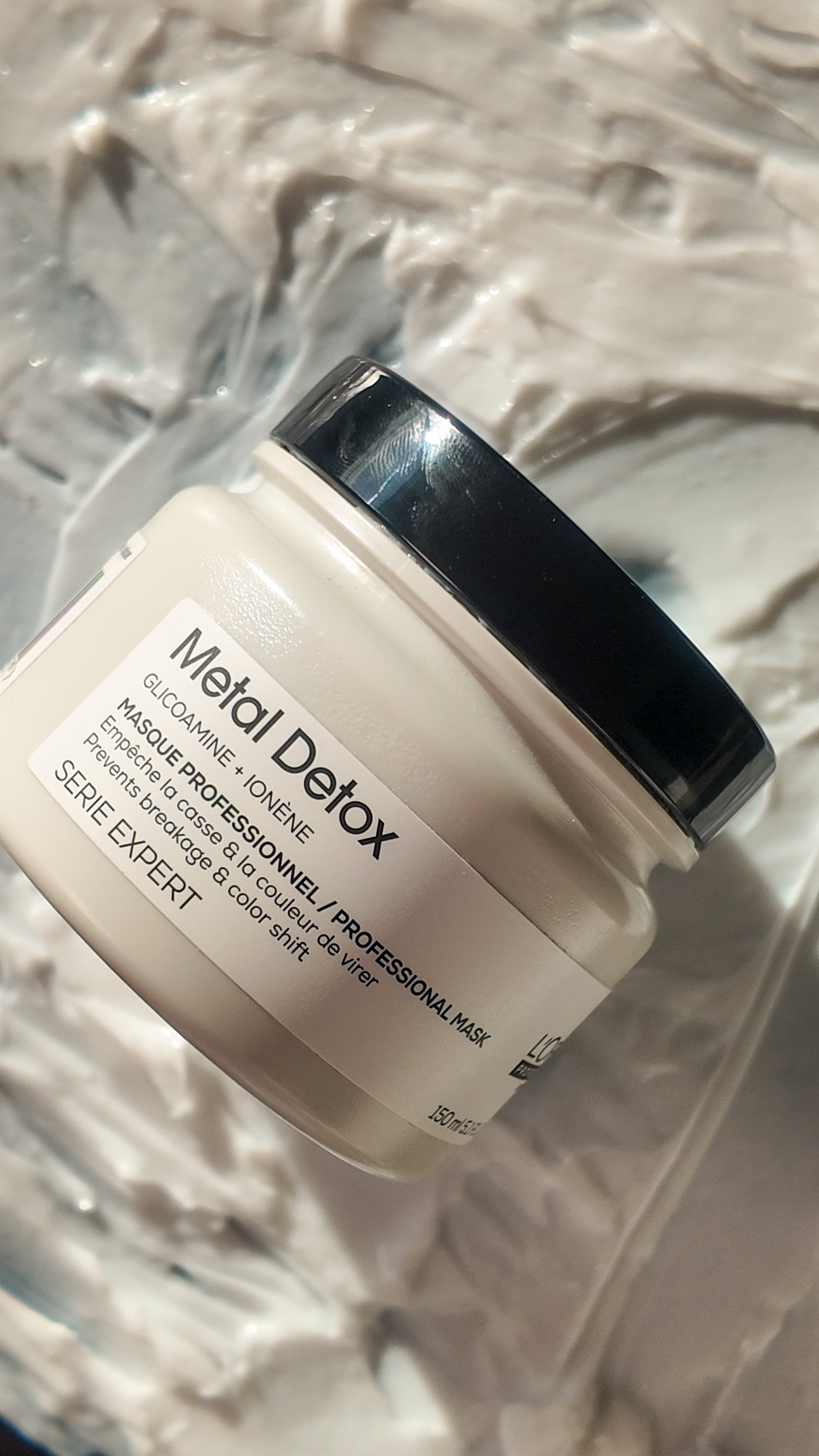 A jar of metal detox facial mask with a black lid, placed on a crinkled silver foil background.