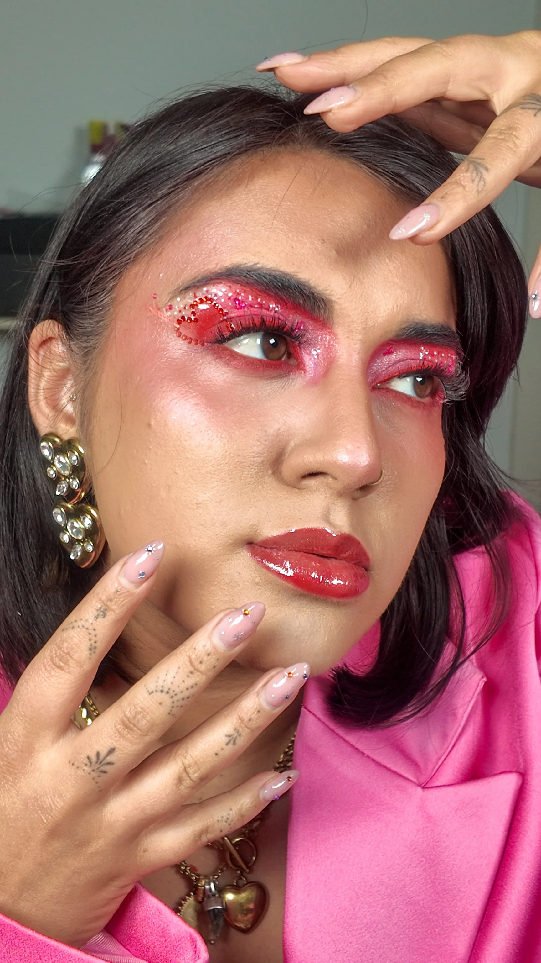 A woman with pink eye makeup, red lipstick, and decorative jewelry, posing with her hand on her face and fingers near her lips.
