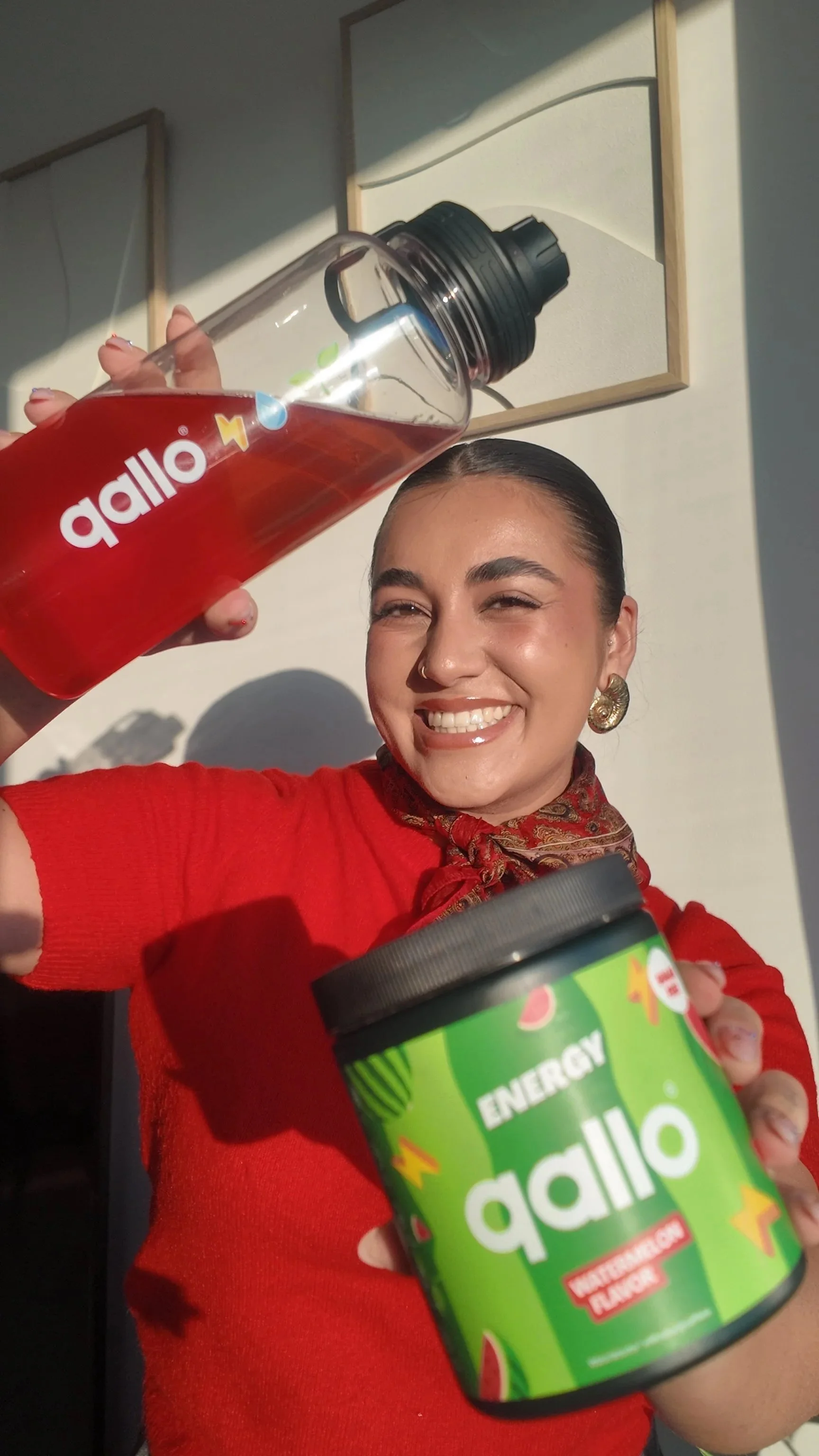 A woman with dark hair tied back, smiling and holding a clear water bottle with a black lid and red liquid, and a green container labeled 'Agli Energy' with a watermelon flavor. She is wearing a red top and a patterned scarf around her neck, standing outside with sunlight illuminating her face.
