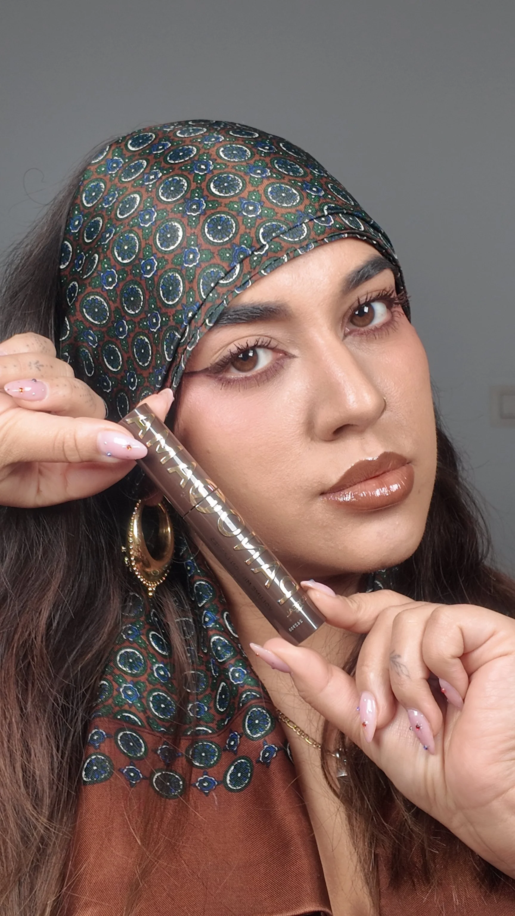 Woman with brown hair and earrings holds a tube of lipstick labeled 'FENTY BEAUTY' while looking at the camera. She wears a patterned headscarf and nude lipstick.