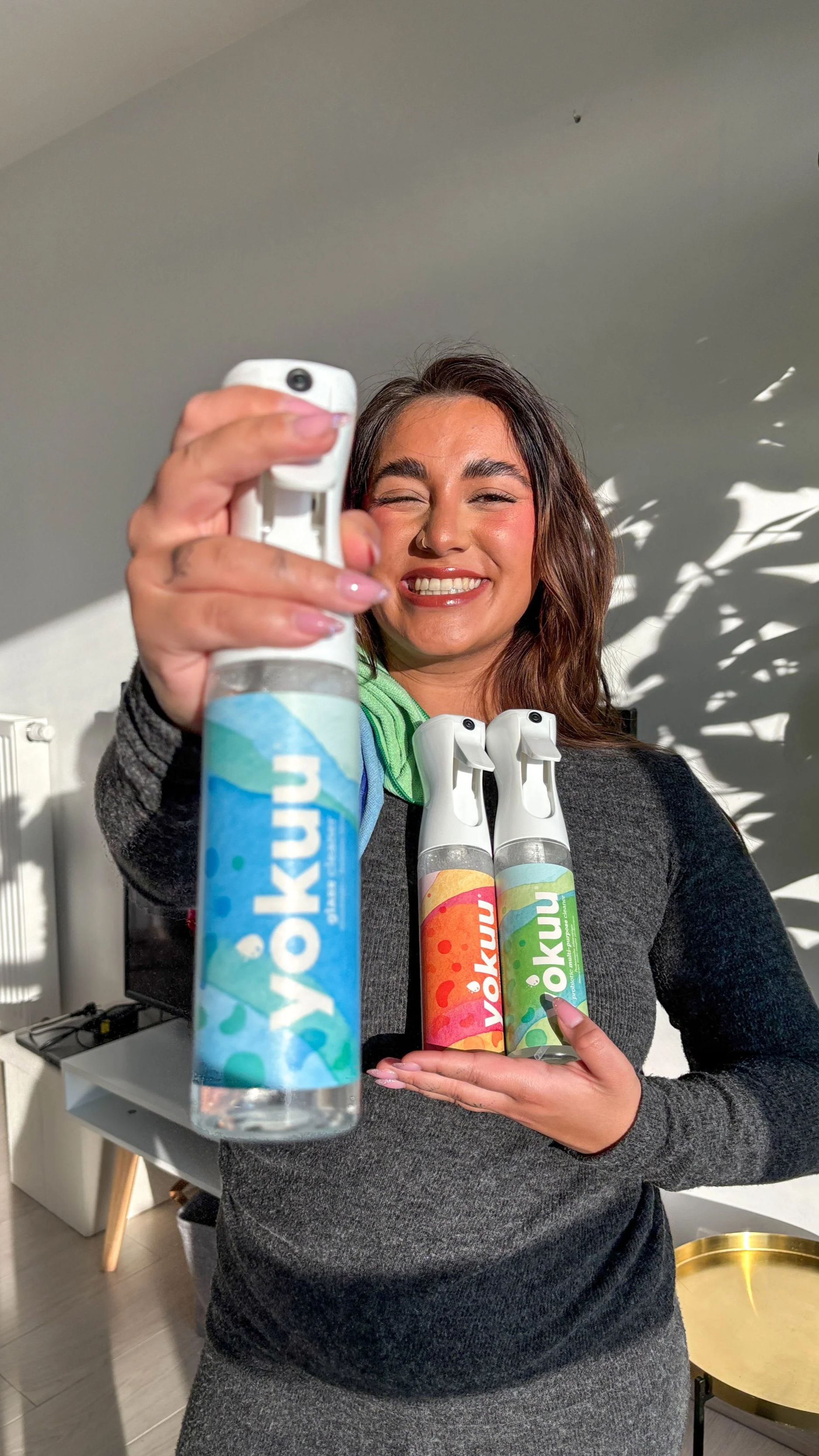 A woman smiling and taking a selfie while holding three bottles of yuknu spray in a room with sunlight and shadow.