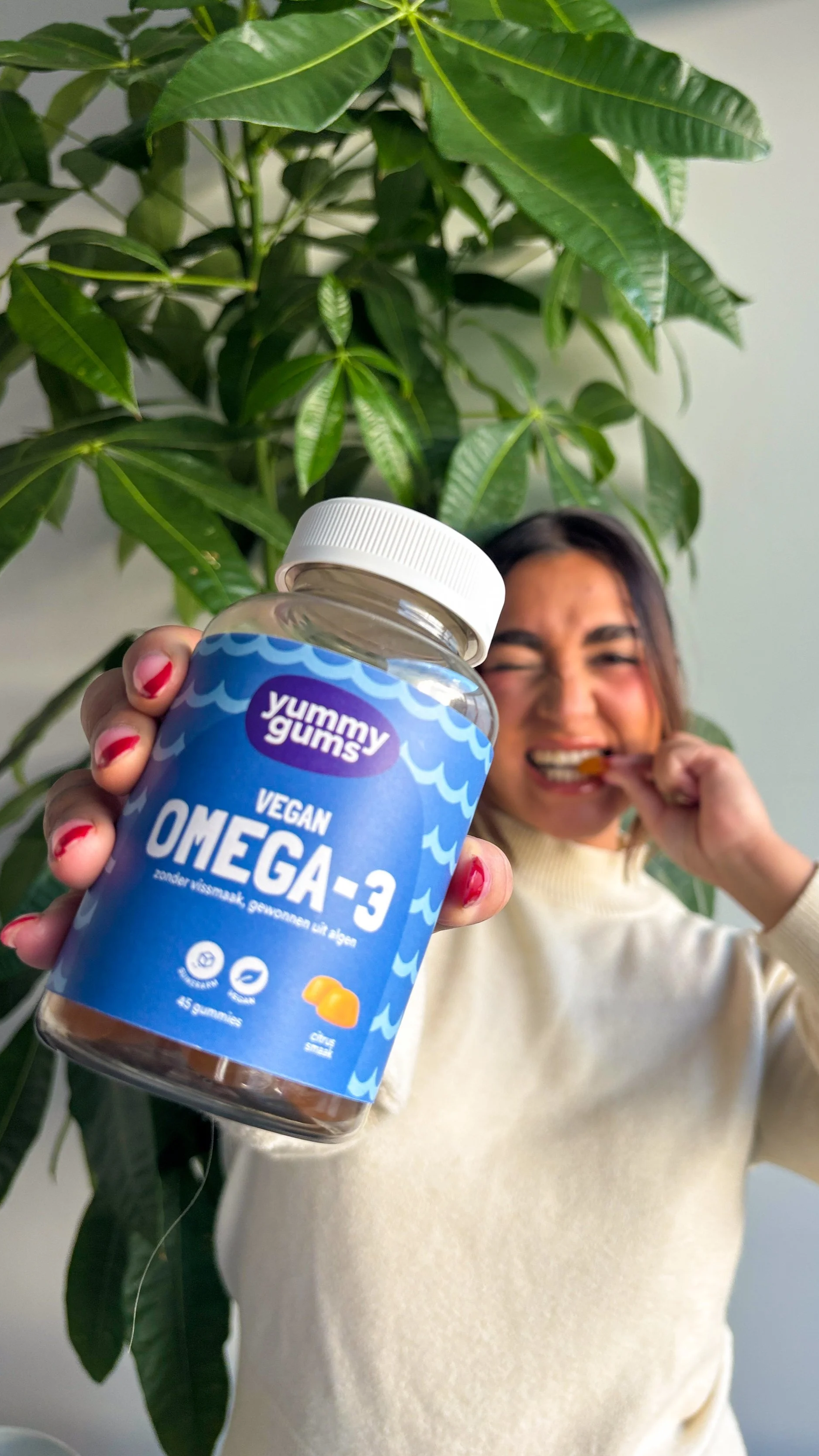 A woman holding a bottle of Yummy Gums vegan Omega-3 gummies in front of a large green plant, with the woman smiling and biting into a gummy.