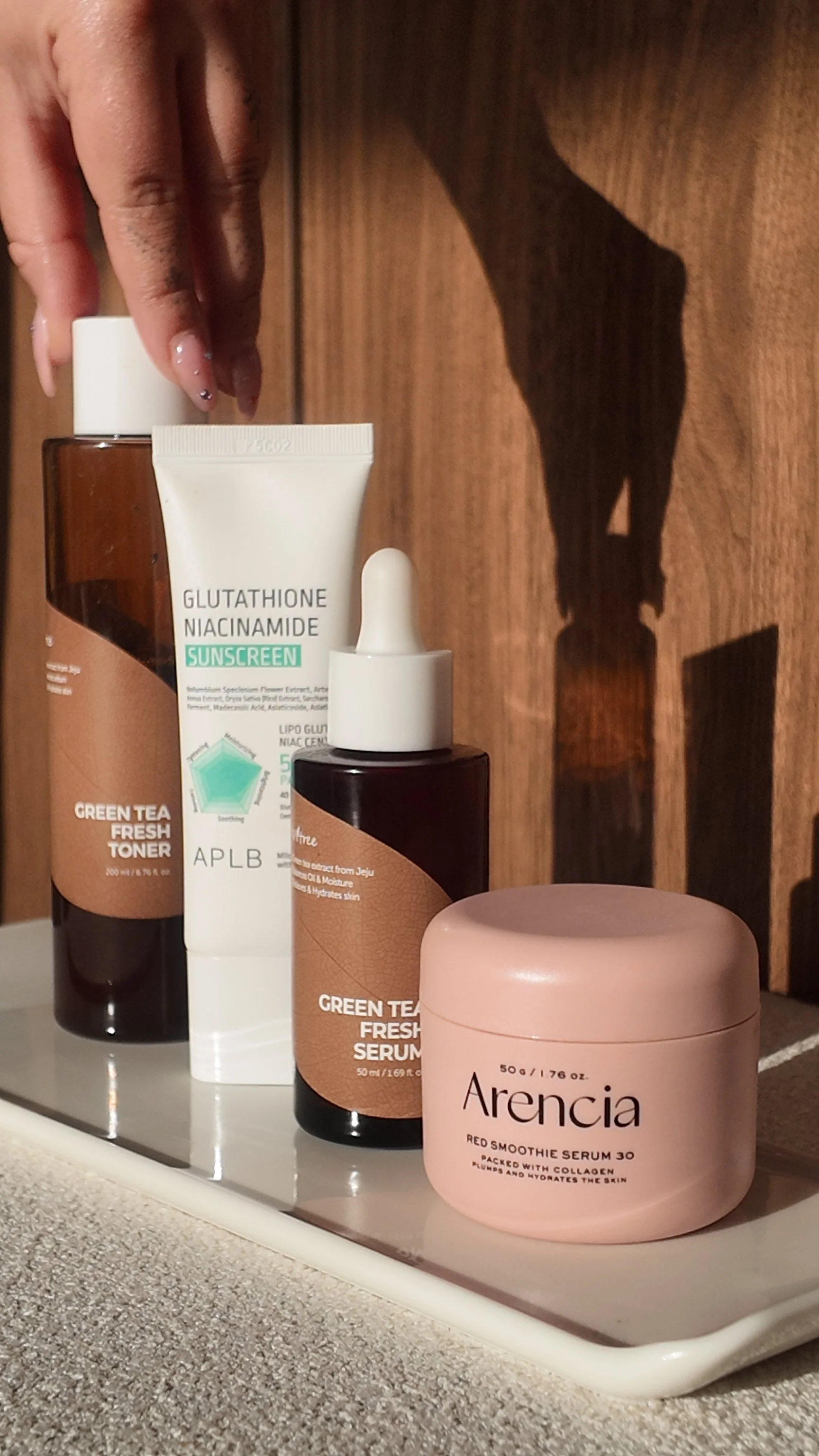 Collection of skincare products including Green Tea Fresh Toner, Glutathione Niacinamide Sunscreen, Green Tea Fresh Serum, and Arencia Red Smoothie Serum placed on a tray against a wooden background.