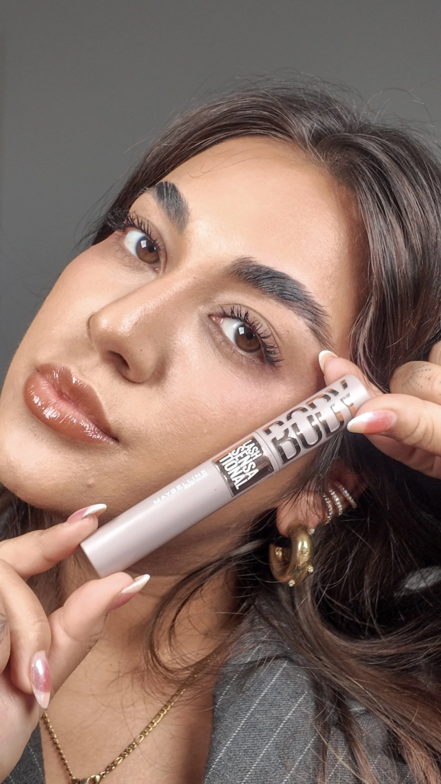 A woman with brown hair, wearing gold earrings, a gold necklace, and a gray pinstripe jacket, holds a tube of Maybelline Lash Sensational Boost mascara near her face. She has makeup on, including eye makeup and glossy lips.