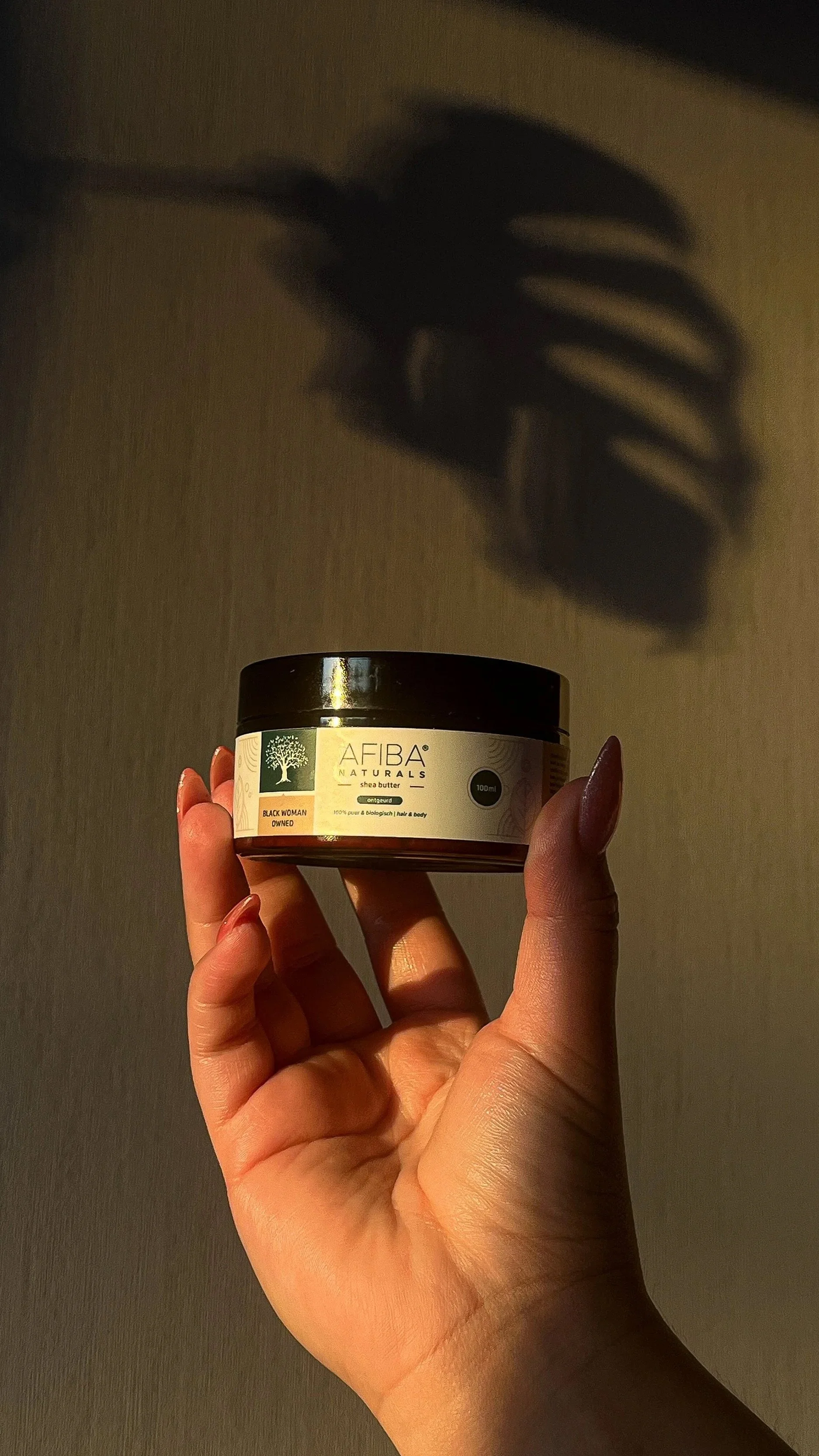 A hand holding a jar of AFIBA Naturals shea butter, with the shadow of the jar cast on a wall behind.
