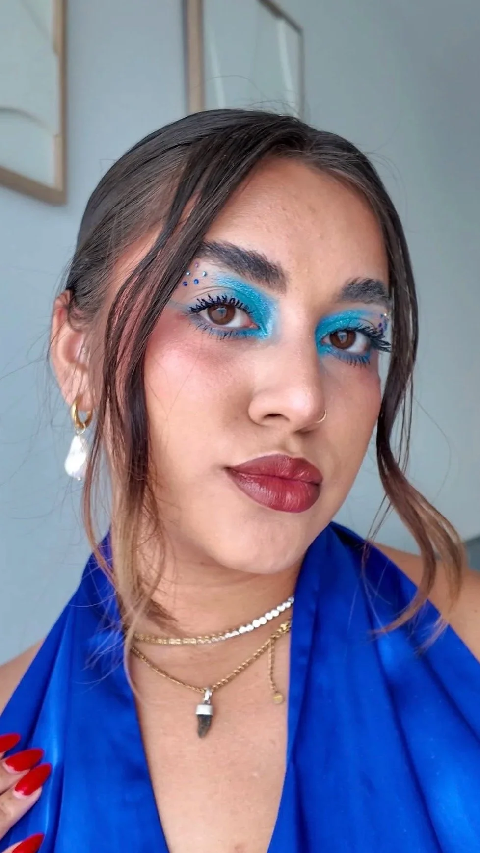 A woman with brown hair and brown eyes wearing bold blue eye makeup, gold hoop earrings, layered necklaces, and a blue halter top.