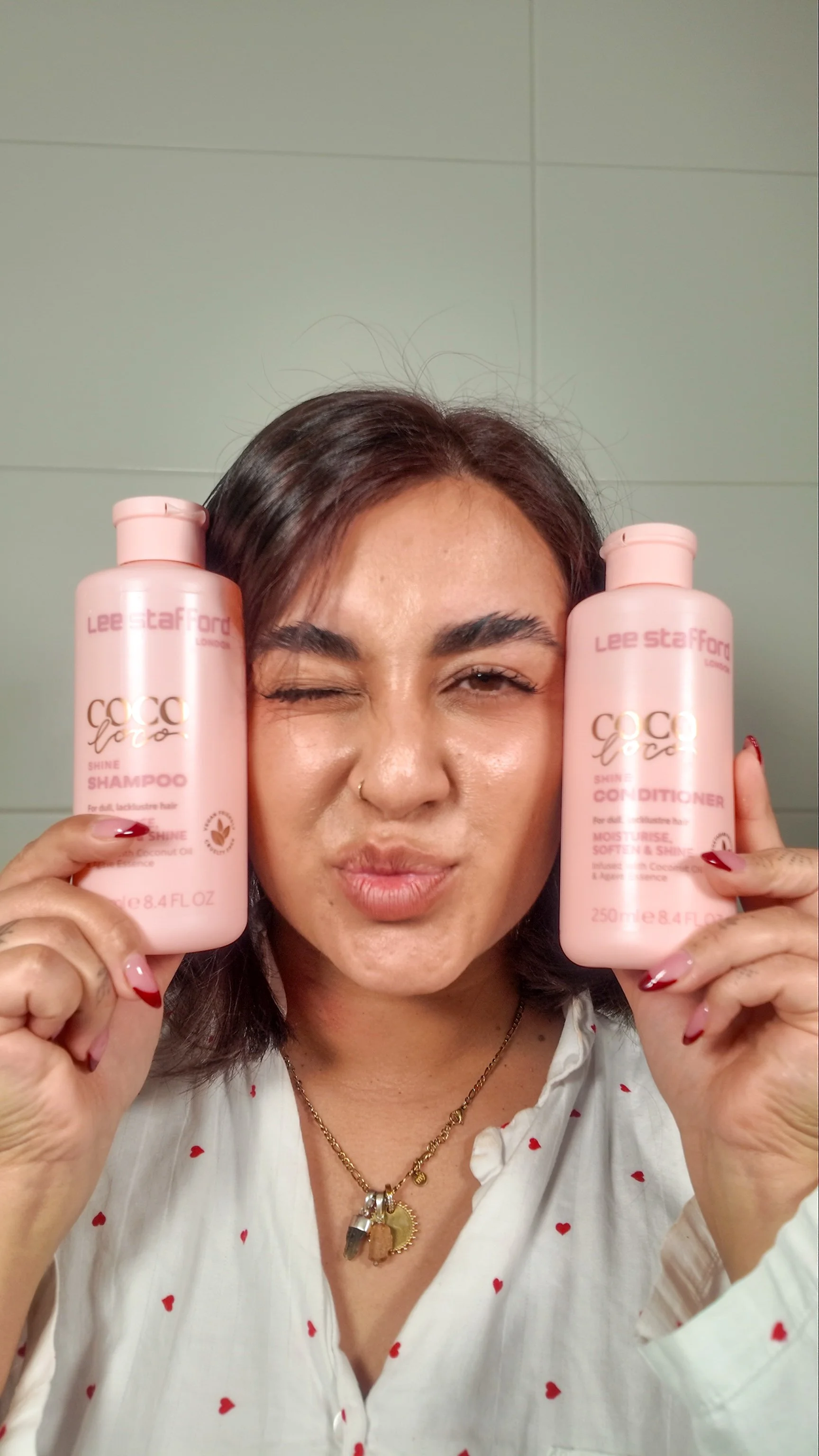 A woman holding up two pink bottles of Lee Stafford Coco La La Shine Shampoo and Conditioner, winking and puckering her lips.