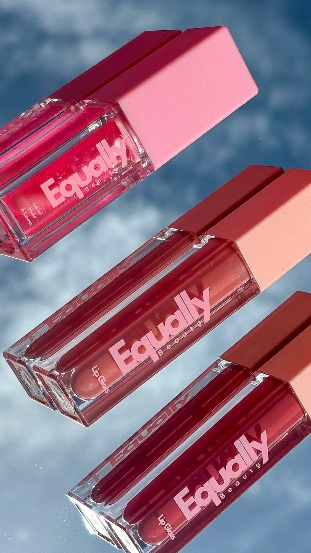 Three lip gloss bottles labeled 'Equaly Beauty' with pink caps, lying on a shiny surface with a blue background.