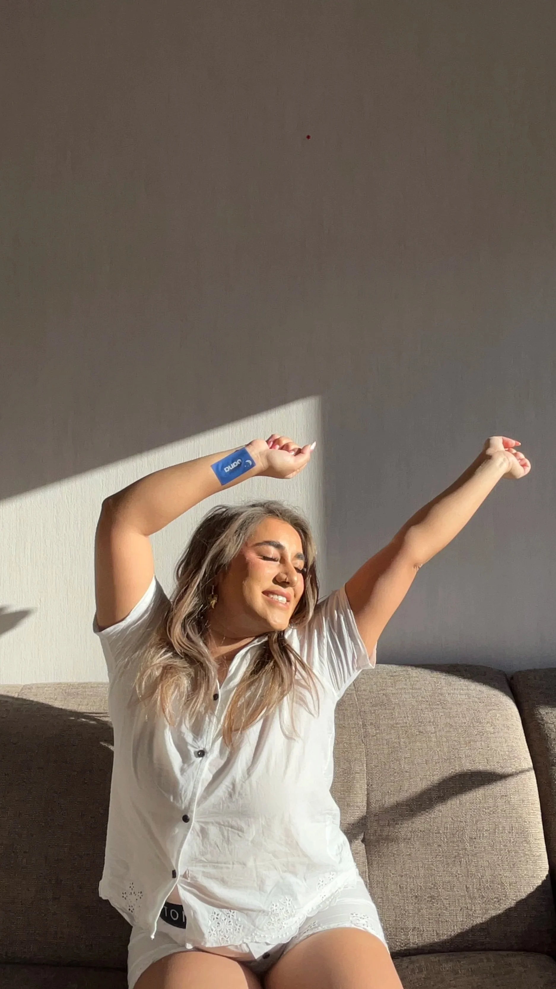 A woman with long, wavy hair sitting on a couch, smiling with eyes closed, stretching with one arm raised above her head and the other bent, wearing a white top and shorts, with sunlight casting shadows on the wall behind her.