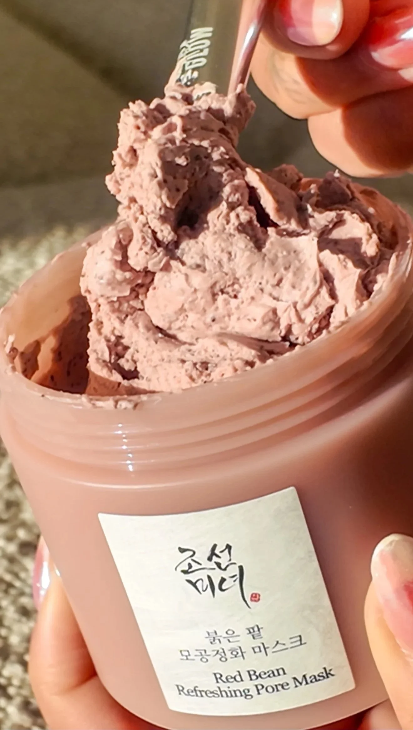 A person holding a container of pink, creamy Red Bean Refreshing Pore Mask with a small spoon. The label on the container has Korean writing and English text.