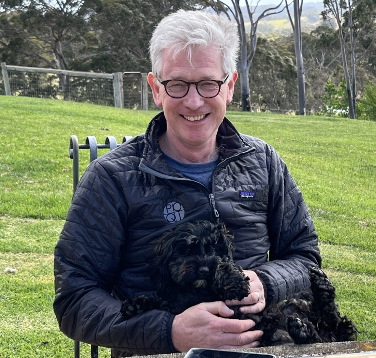 Man smiling while holding a small black dog outdoors on a grassy area.