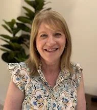 Smiling woman with blonde hair in a patterned blouse, indoor setting with a plant in the background.