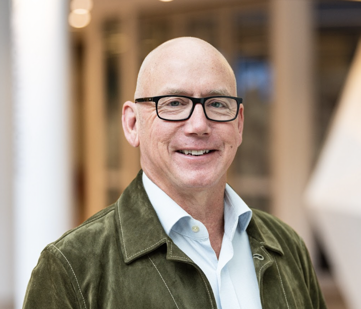 Smiling man with glasses and a green jacket in an indoor setting.