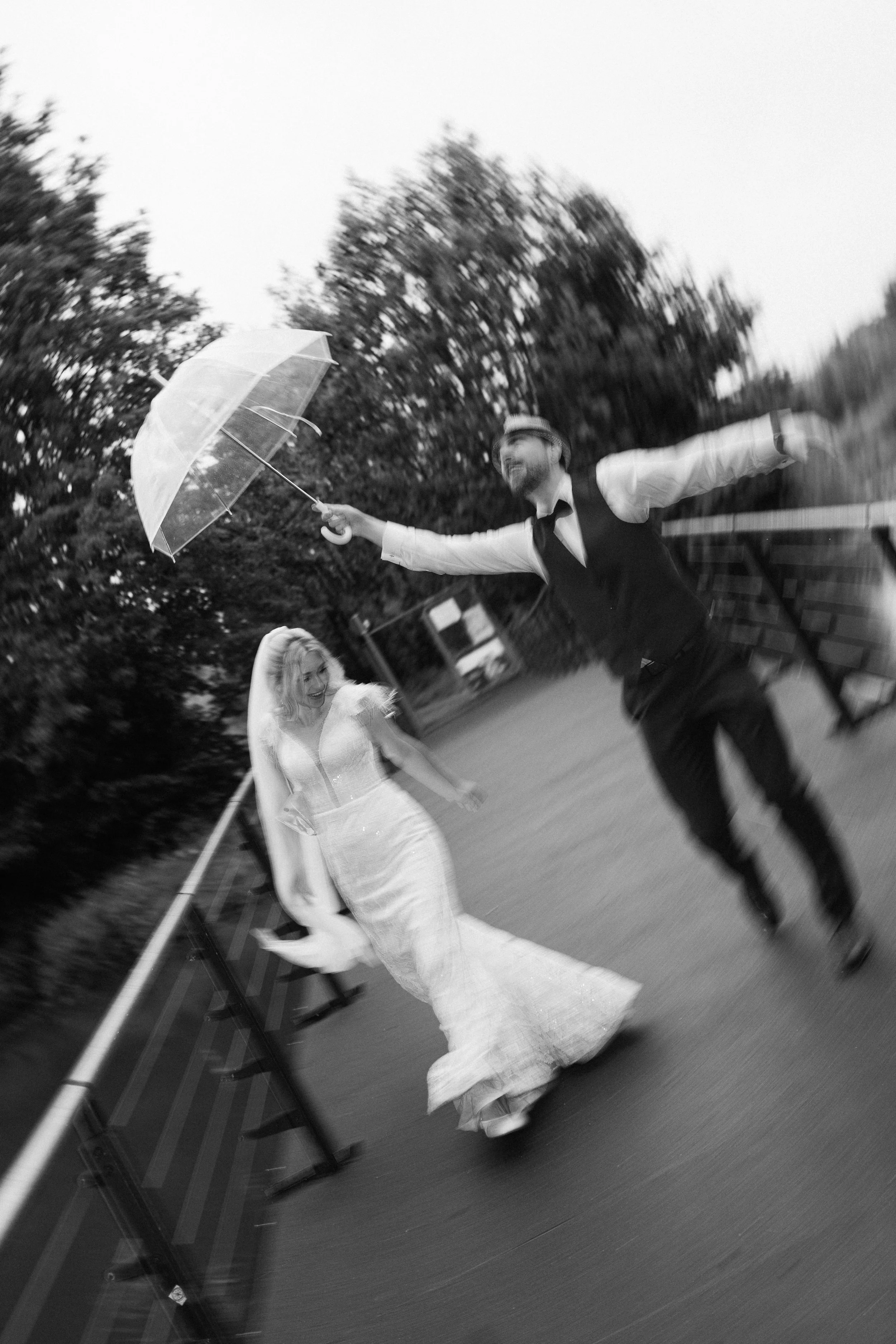 Ein Mann in Anzug und Krawatte mit Hut hält einen transparenten Regenschirm und umarmt eine Frau im Hochzeitskleid, beide lachen.