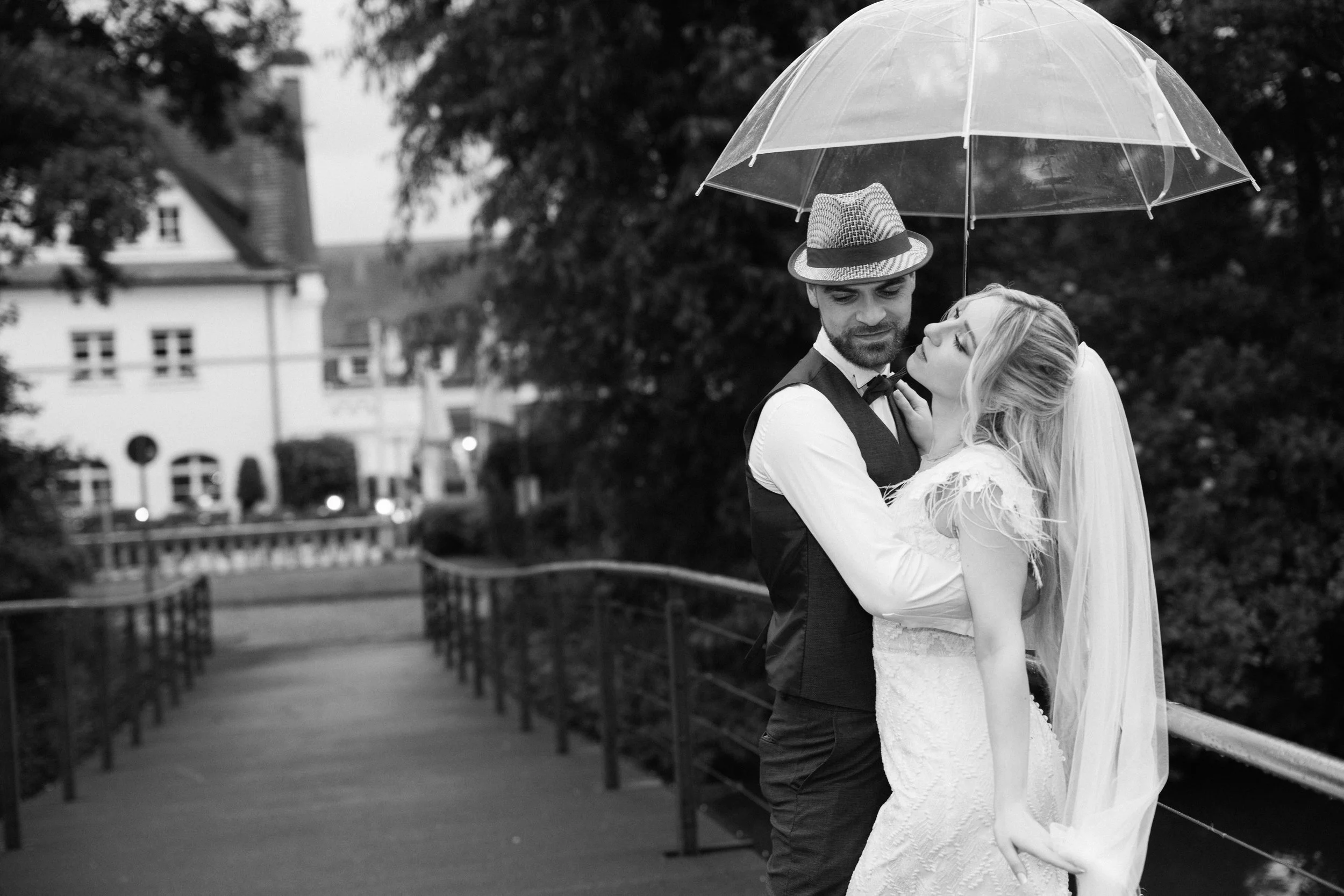 Ein Mann und eine Frau, wahrscheinlich Braut und Bräutigam, stehen sich nahe beieinander unter einem transparenten Regenschirm im Regen. Sie schauen sich liebevoll an, in einer urbanen Umgebung mit Bäumen, Häusern und einem Gehweg im Hintergrund, in 